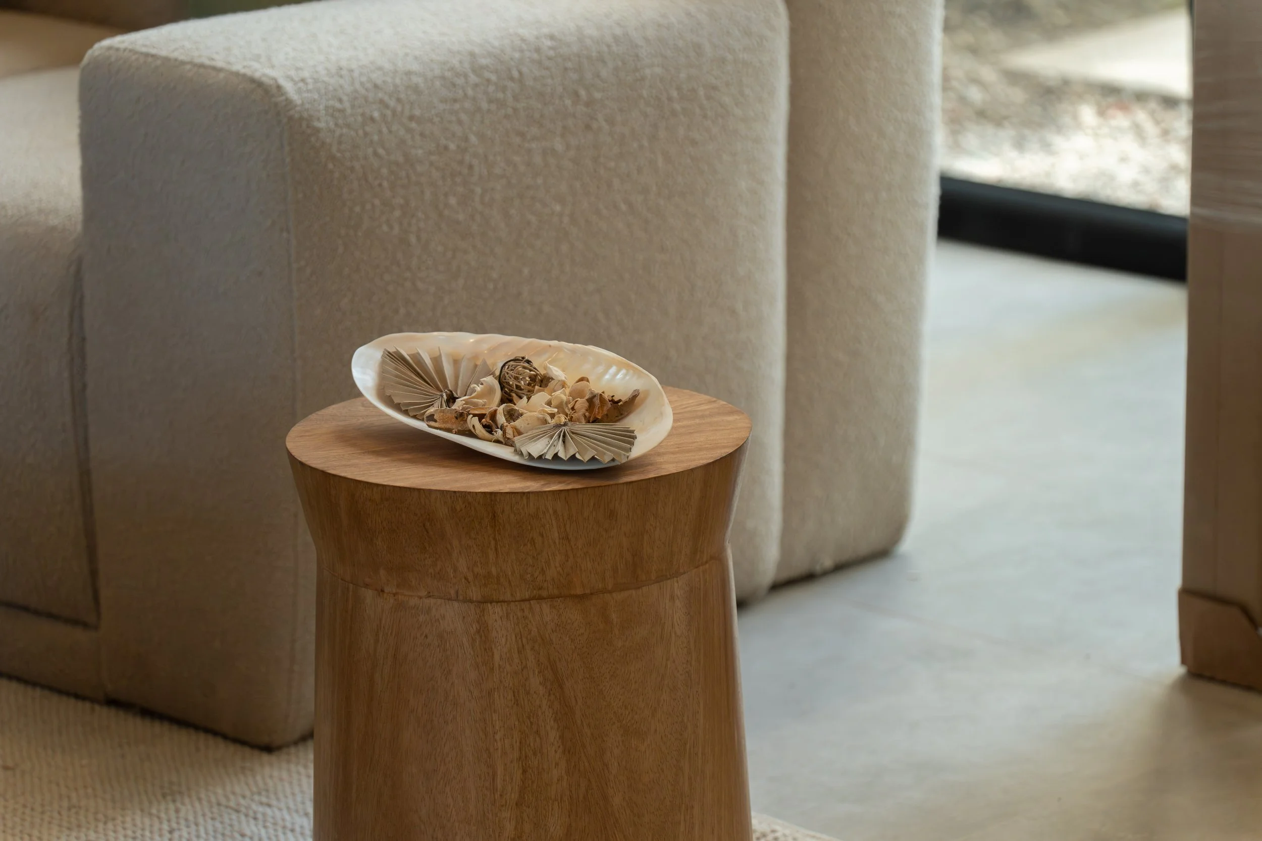 Close-up of a wooden side table with a shell-shaped dish containing dried flower petals and decorative accents, placed next to a beige upholstered sofa.
