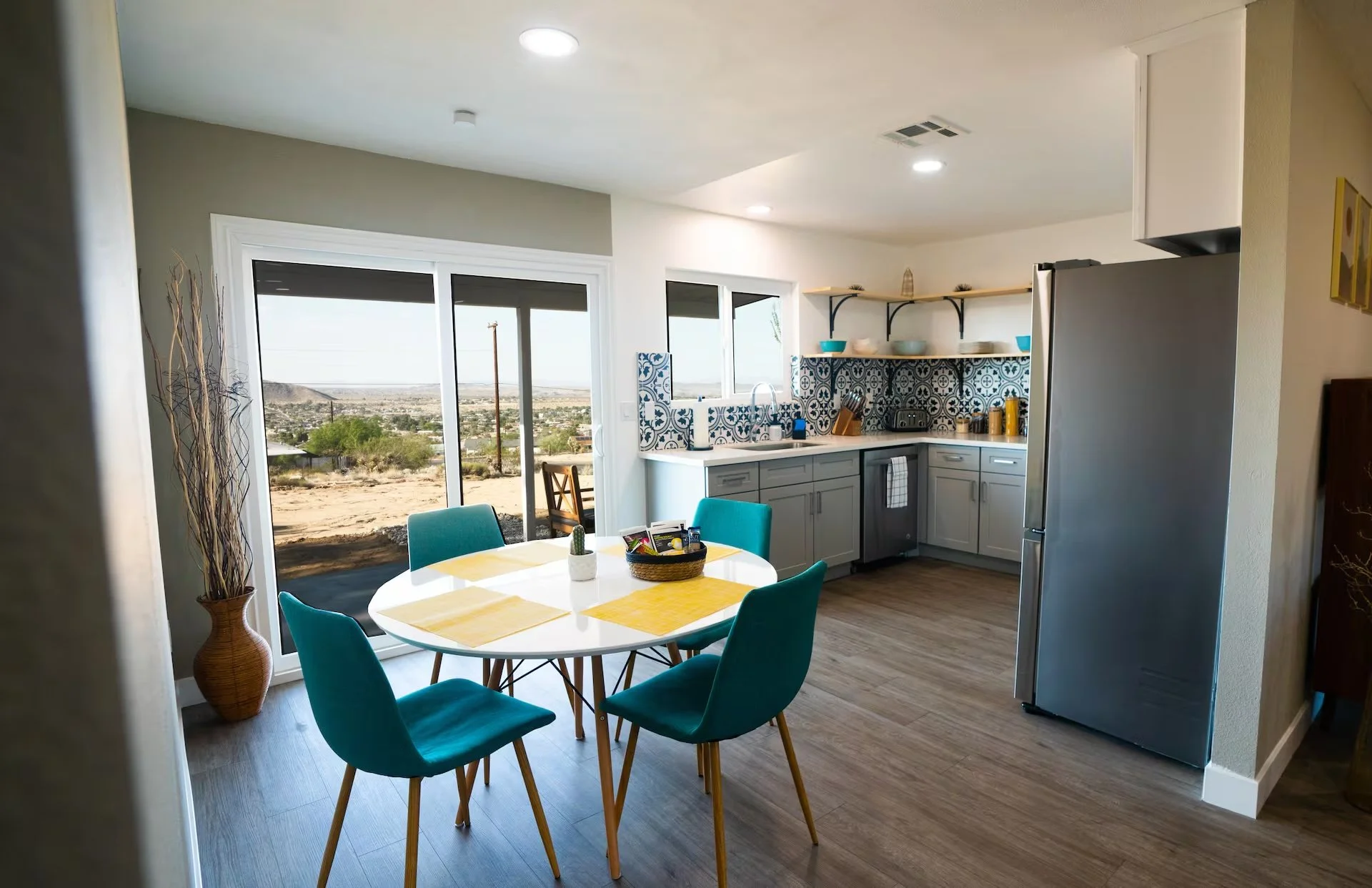 Modern kitchen and dining area with sliding glass door leading to an outdoor desert landscape, teal dining chairs around a white table with yellow placemats, blue patterned backsplash, open shelves with bowls and cups, stainless steel refrigerator, a