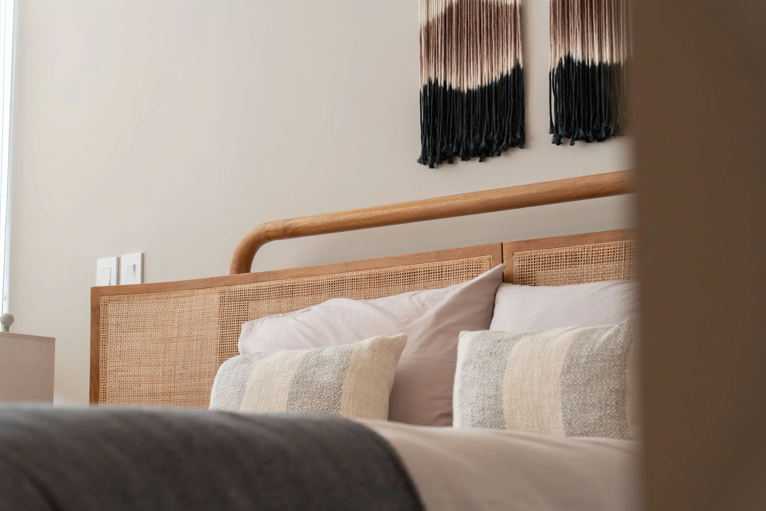 Close-up view of a bed with beige and white patterned pillows, a wooden headboard, and a decorative wall hanging with black and pinkish-beige strands.