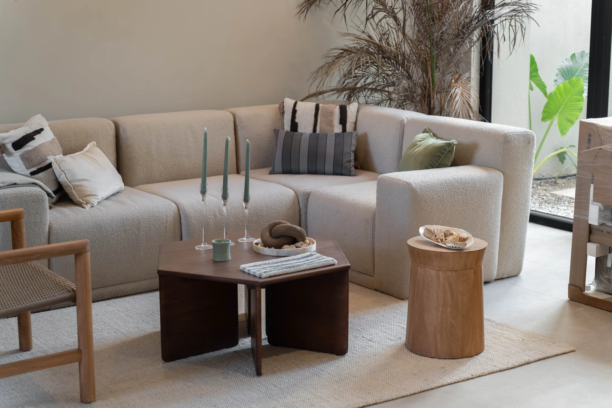 Living room with a beige sofa decorated with various pillows, a wooden coffee table with candles and decorative objects, a large indoor plant, and a side table with a bowl of shells, near a window showing outdoor greenery.