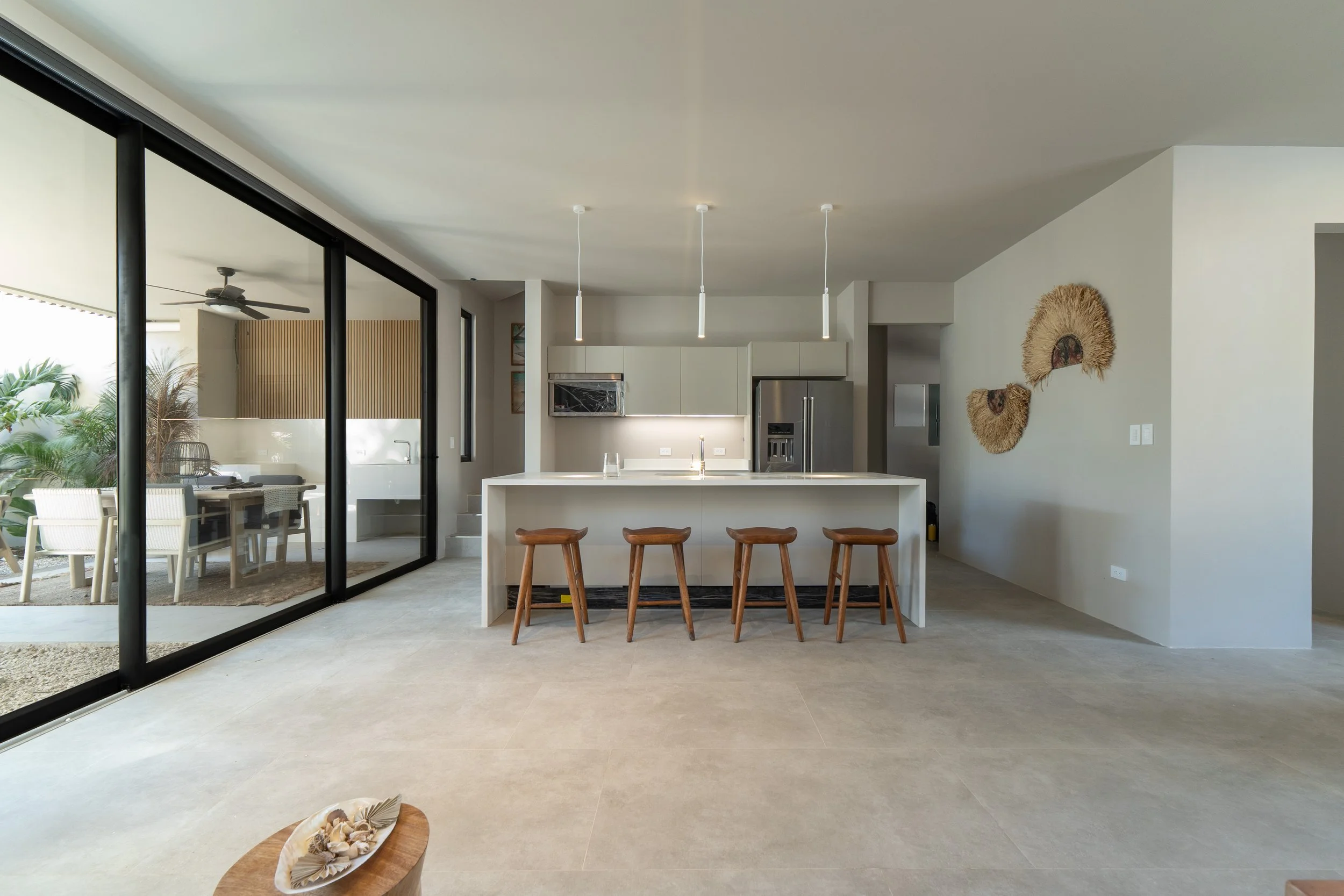 Modern open-concept kitchen and dining area with large sliding glass doors leading to an outdoor patio. The kitchen has a white island with four wooden stools, pendant lighting, and built-in stainless steel appliances. The living space includes decor