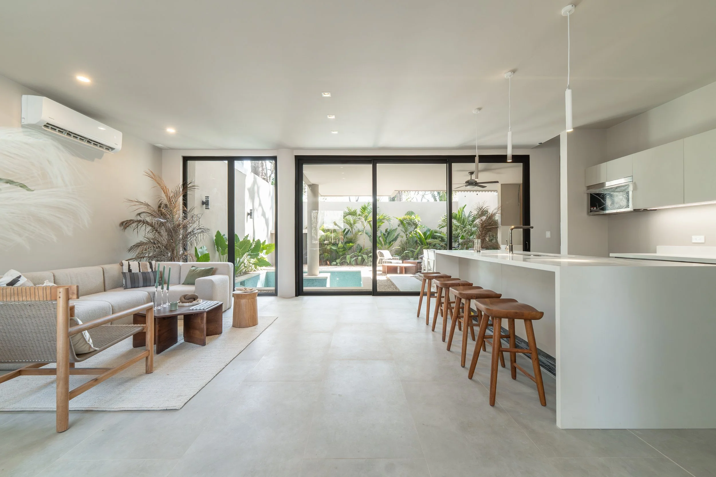 Open-concept living room and kitchen with sliding glass doors leading to a patio and garden visible outside. Interior features include a beige sofa, wooden chairs, a white kitchen island, and indoor plants.