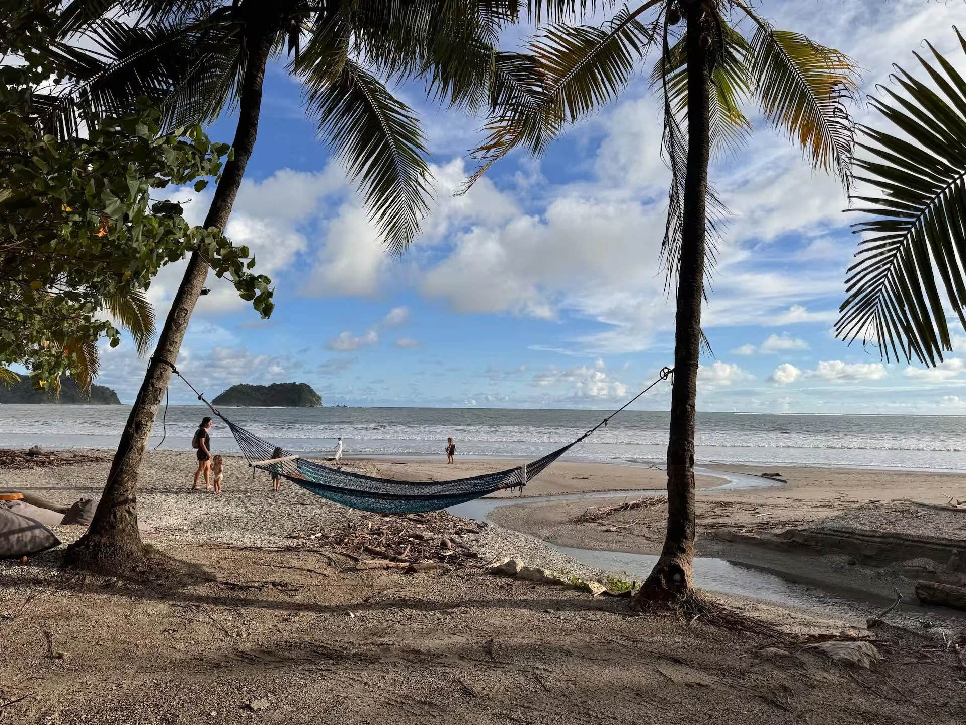 A tropical beach with palm trees, a hammock between two trees, and people walking near the shoreline under a partly cloudy sky.