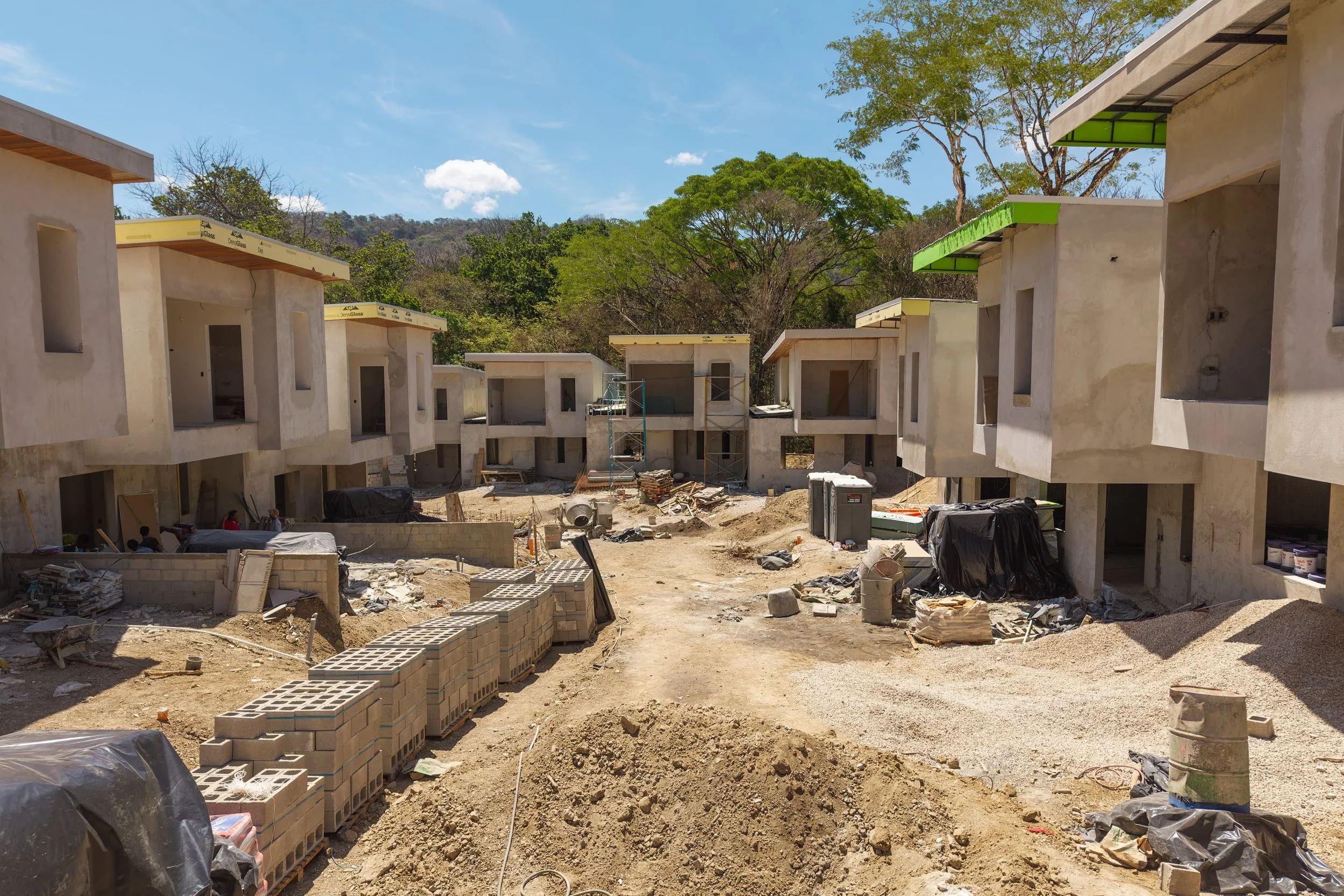 Construction site with unfinished houses, scattered construction materials, and scaffolding, with trees and a blue sky in the background.