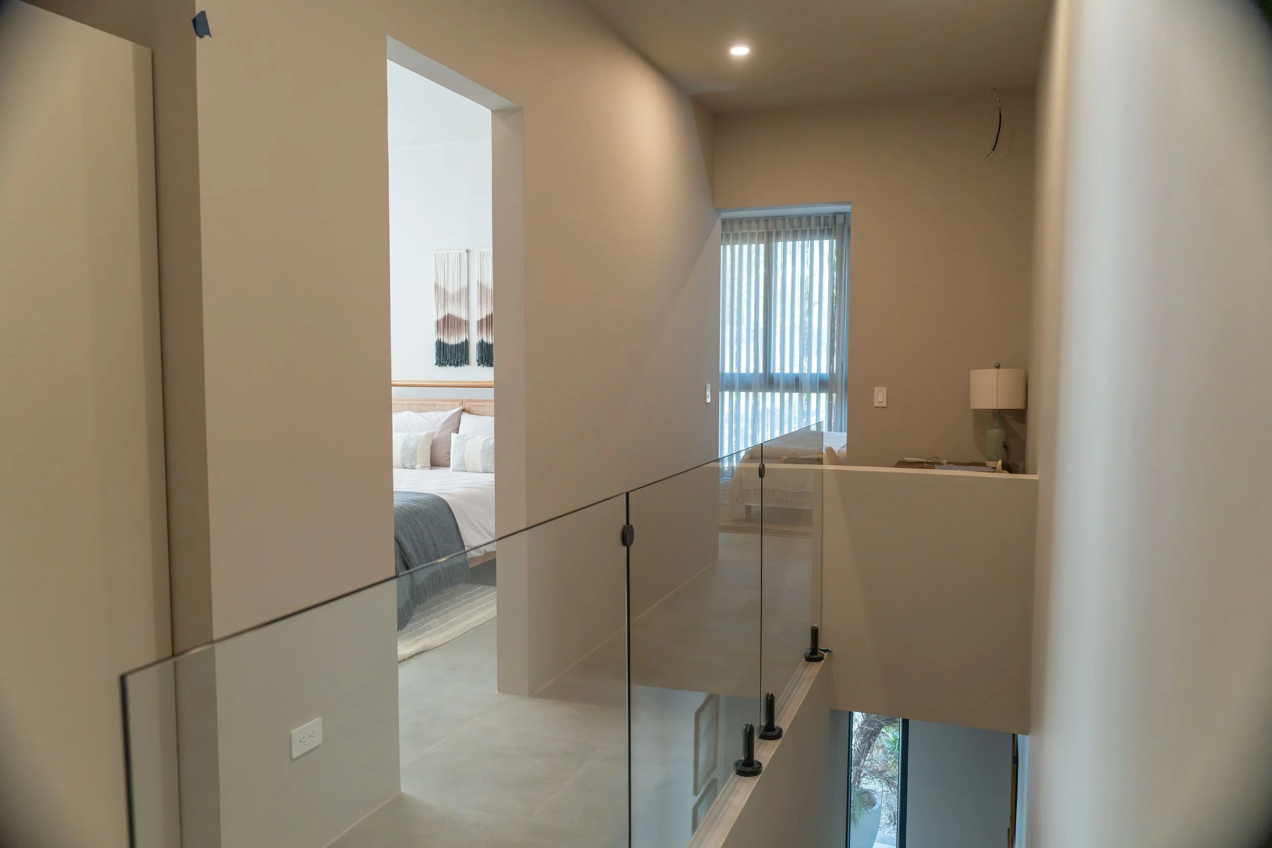 Interior of a modern house with a staircase, a sitting area visible through an open doorway, and large windows with vertical blinds.