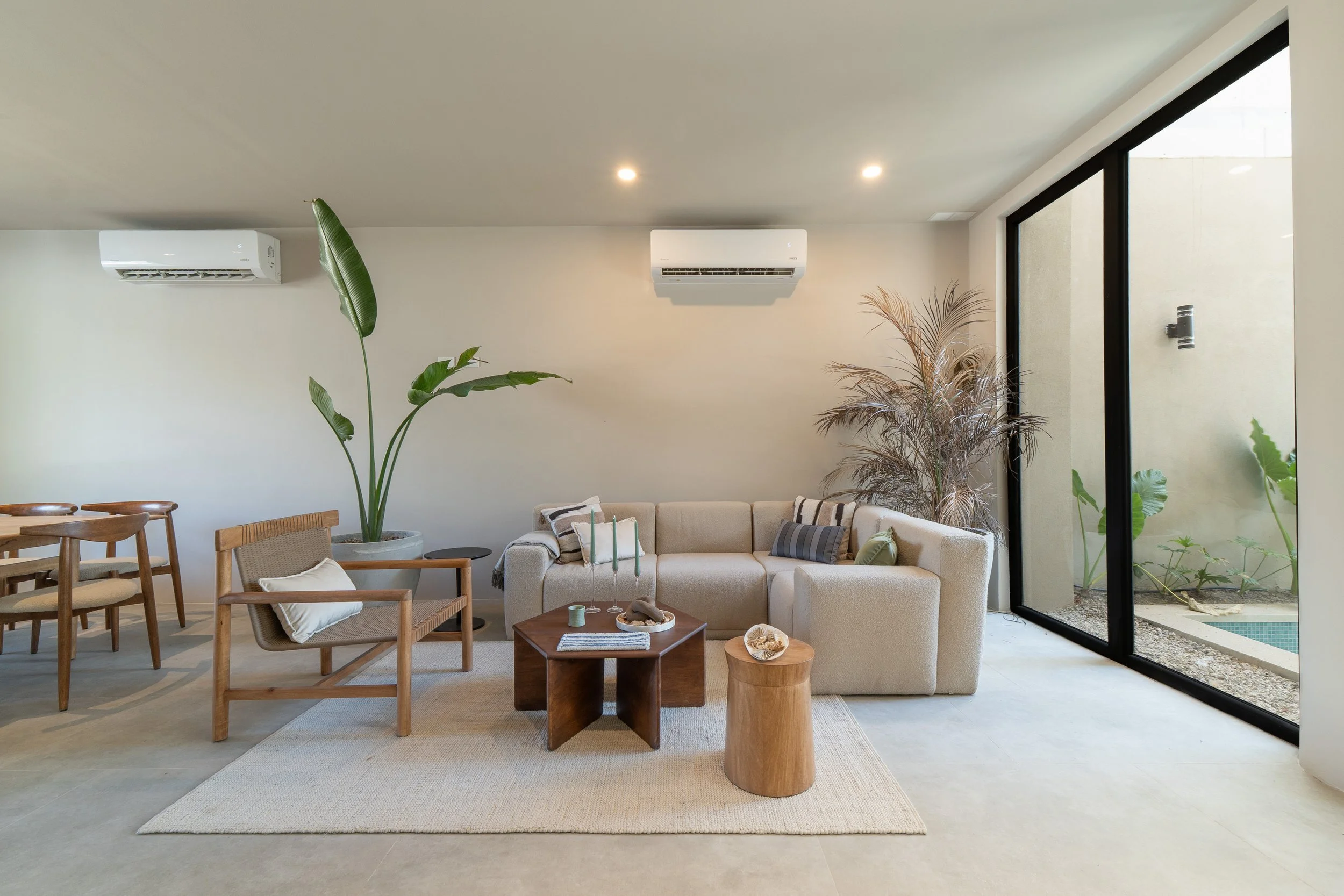 Living room with beige sectional sofa, wooden chairs, and coffee tables, large potted plants, sliding glass door overlooking outdoor area, neutral color palette, minimalist decor.