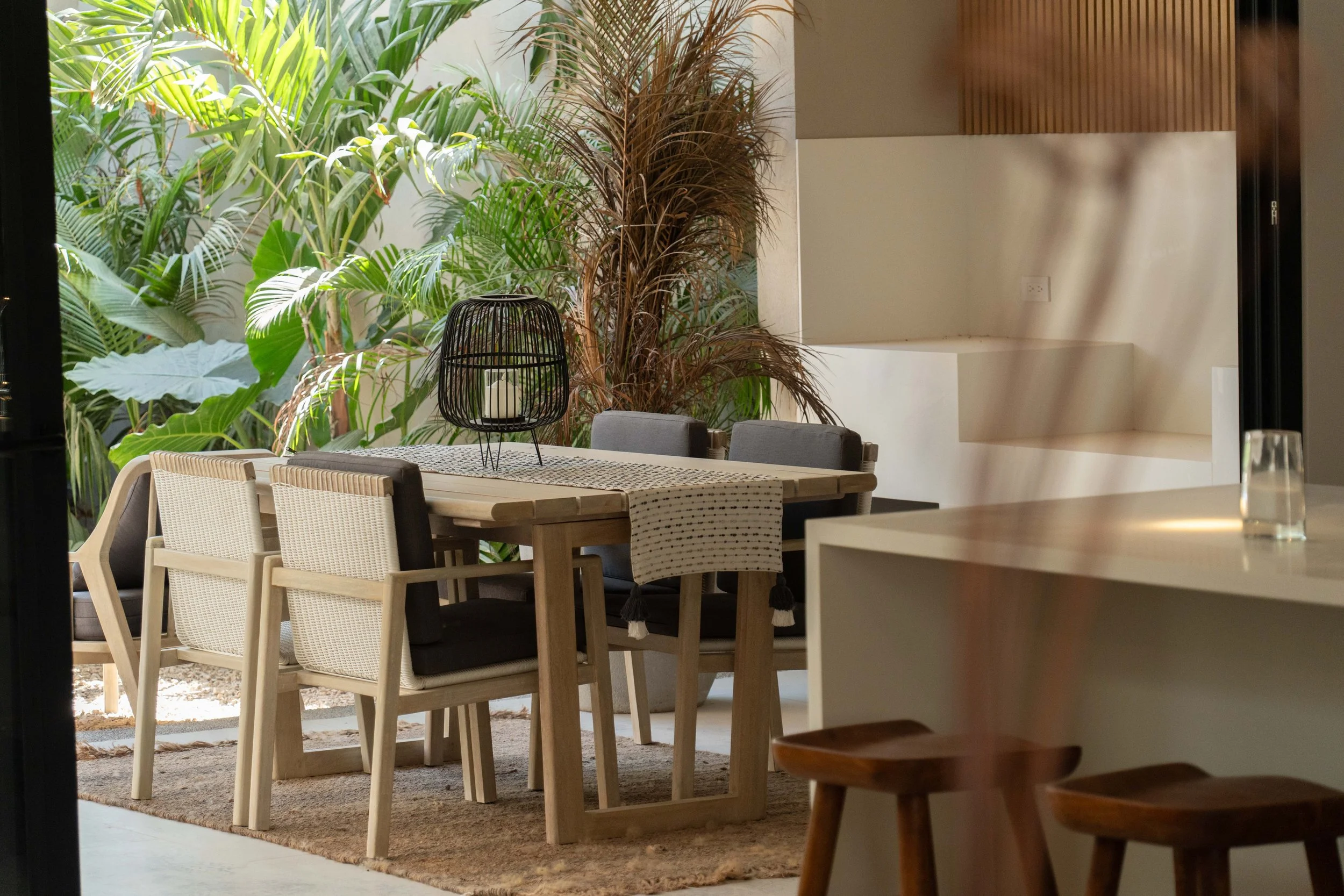 A dining area with a large wooden table, grey chairs, a black lantern with a candle, and lush green tropical plants in the background, with sunlight casting shadows through nearby windows.