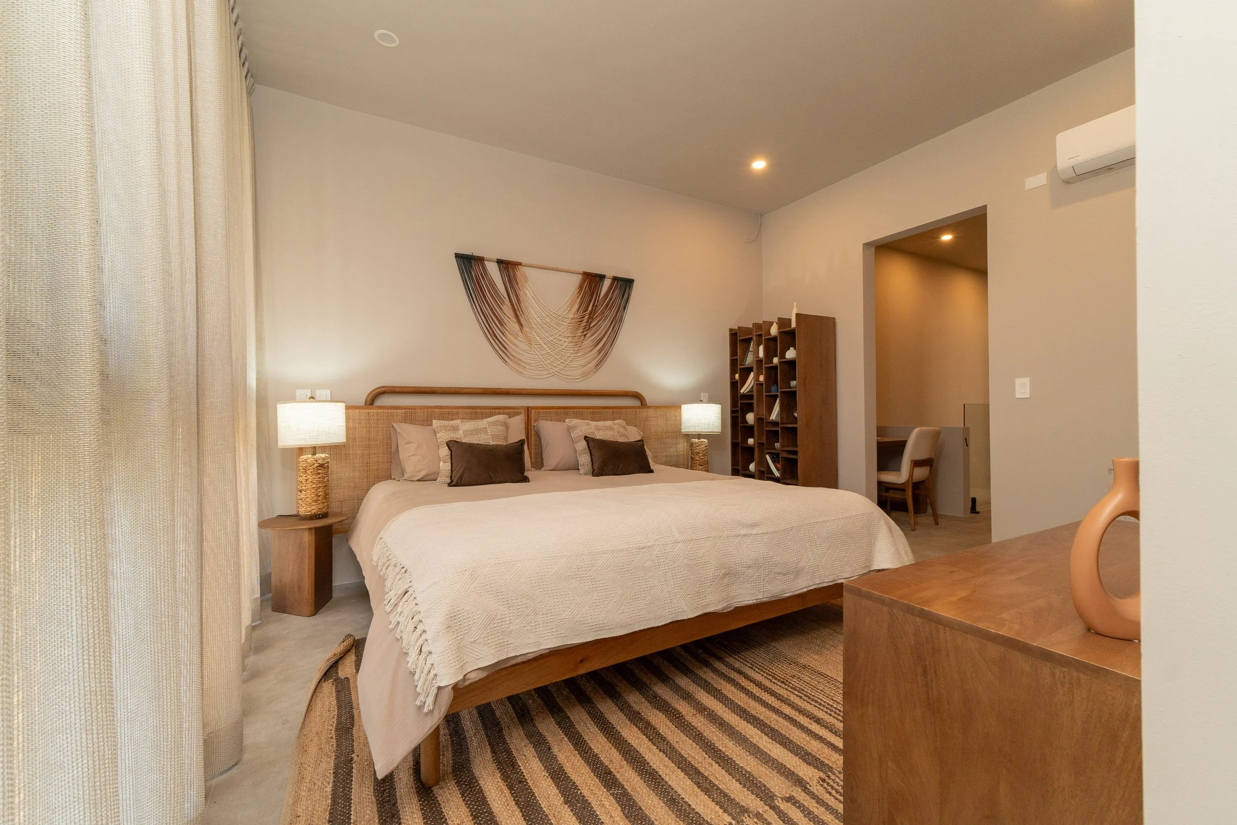Bedroom with a bed, nightstands, two lamps, a woven wall hanging, a bookshelf, and a desk.
