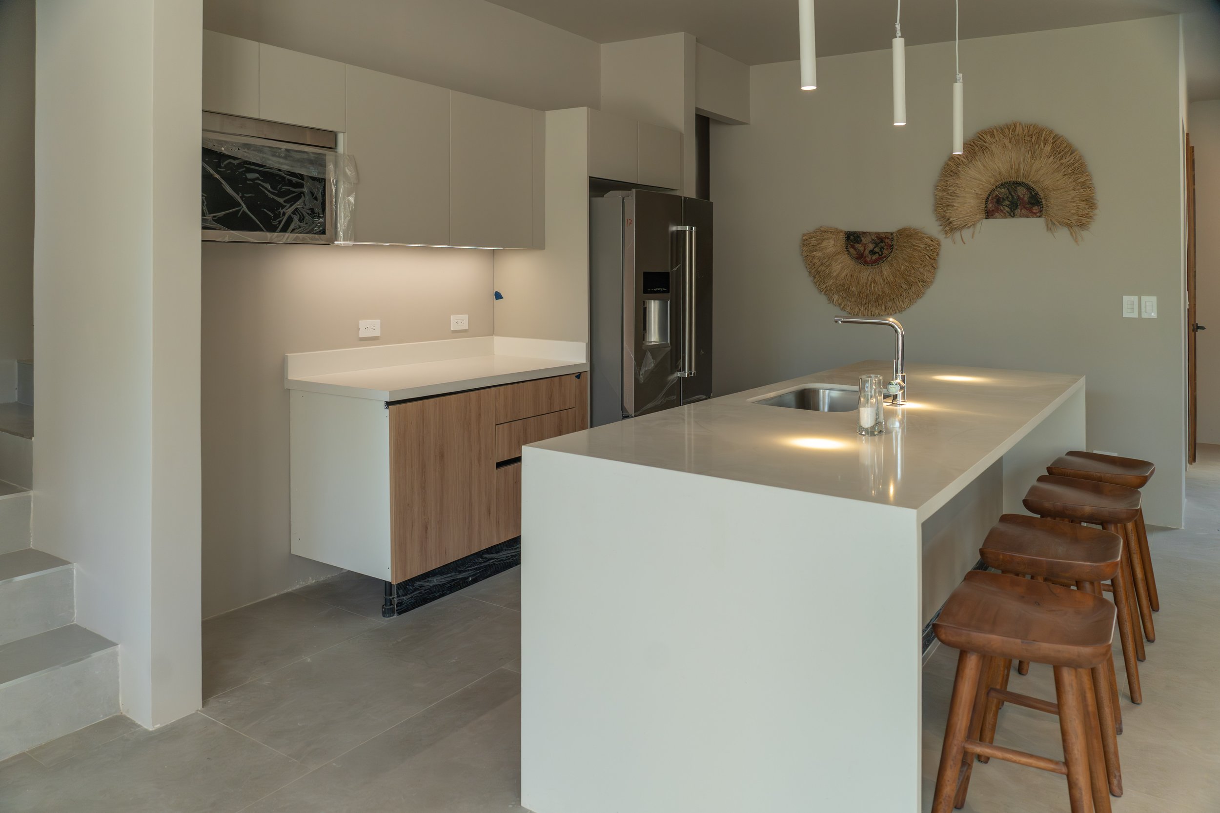 Modern kitchen with beige walls, white countertops, wooden kitchen island with four brown stools, and wall decorations resembling large fans made of straw with circular centers.