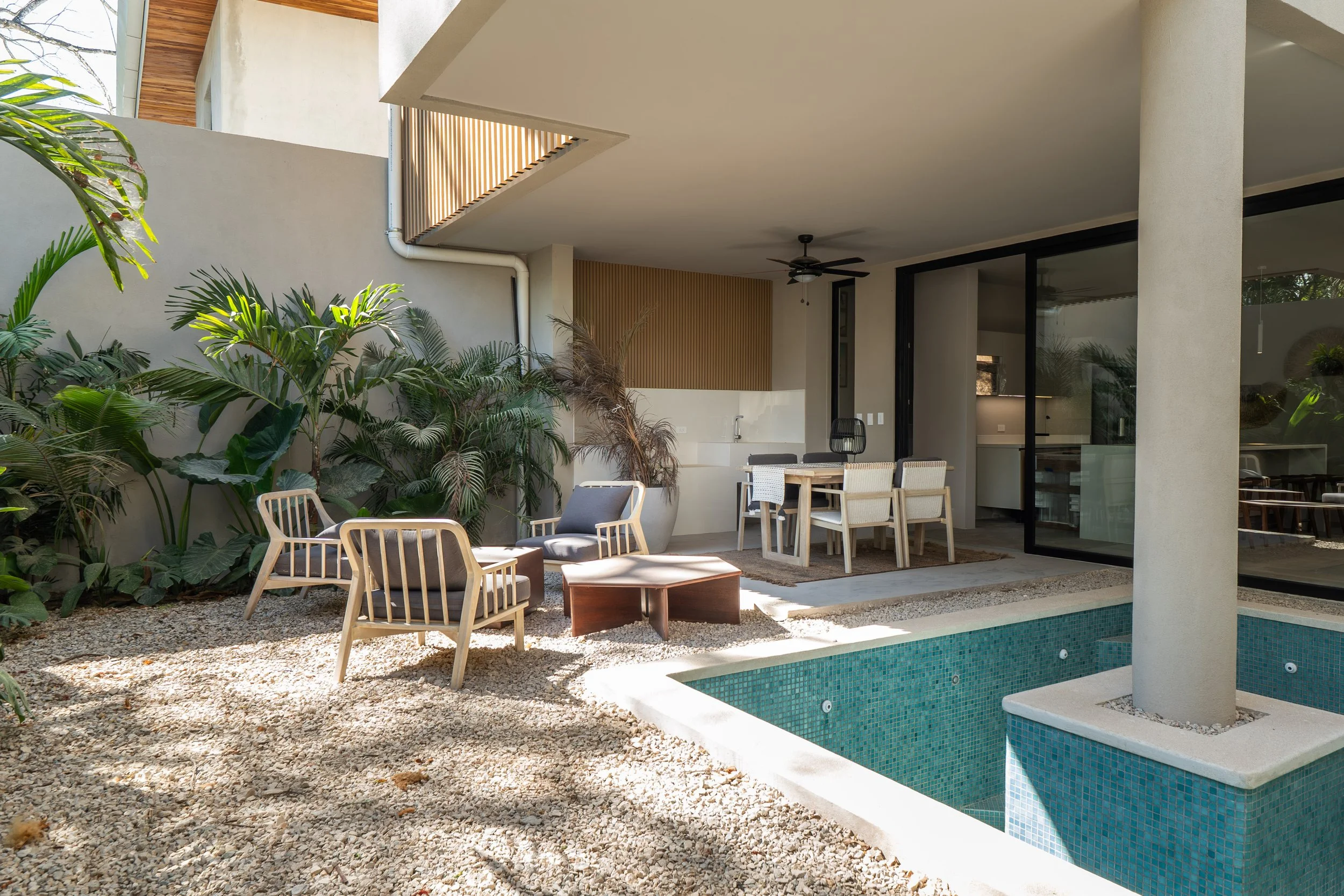 Modern outdoor patio area with a swimming pool, surrounded by garden plants and outdoor furniture including chairs and a dining table under a covered patio with ceiling fan.
