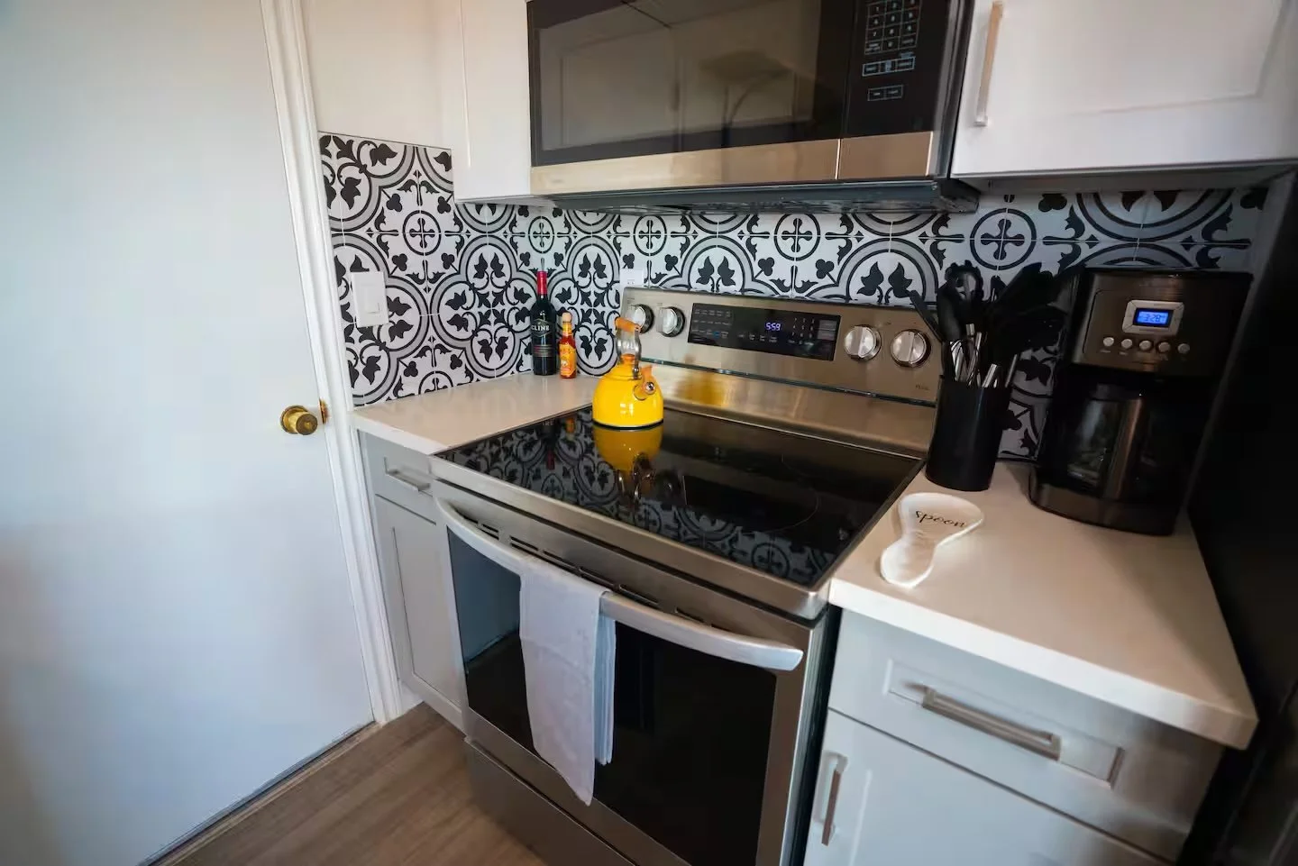 Kitchen with black and white patterned tile backsplash, white cabinets, stainless steel stove, microwave, black coffee maker, and utensils in a black container.