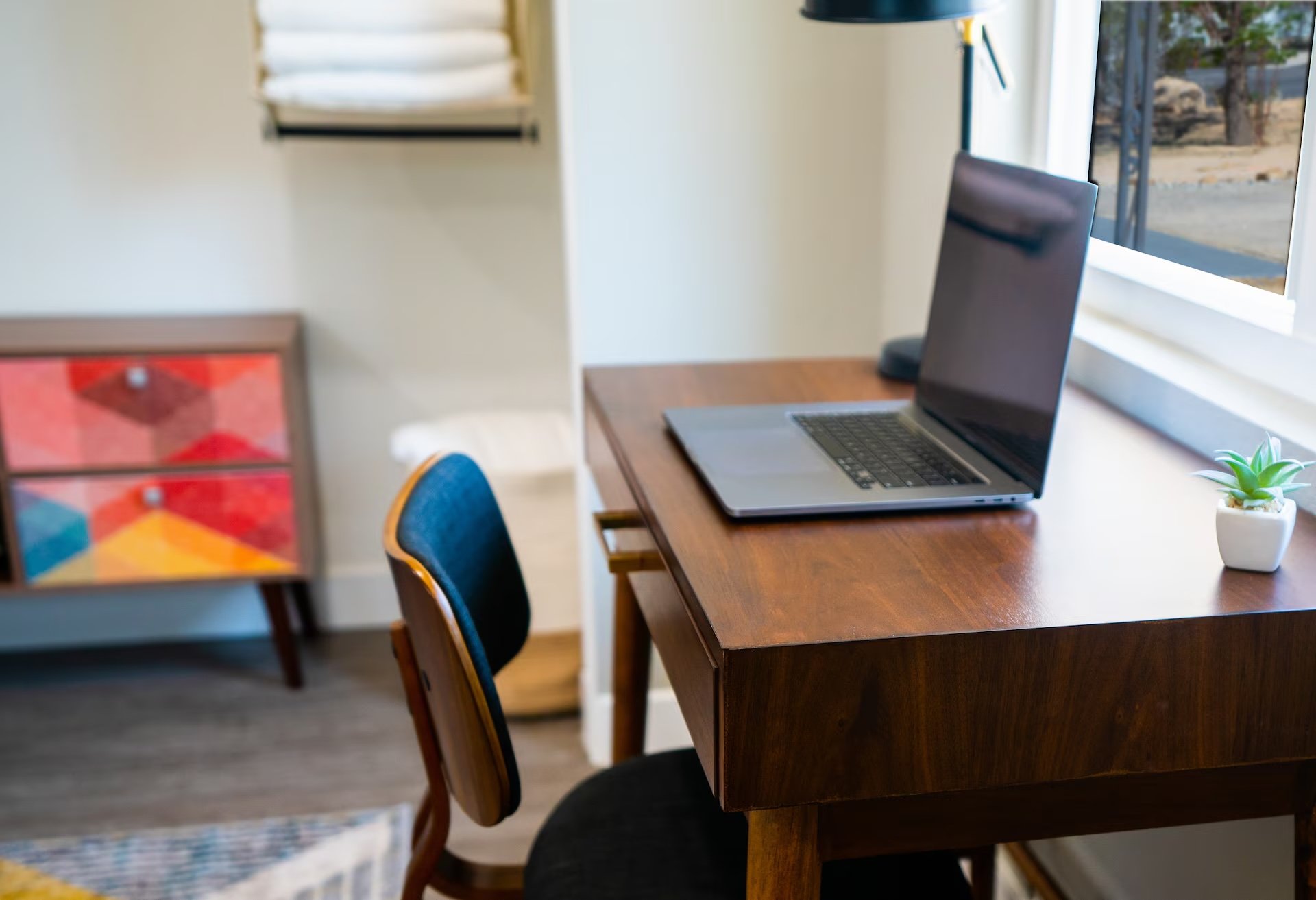Home office with a wooden desk, open laptop, small potted succulent, and a window with outdoor view.