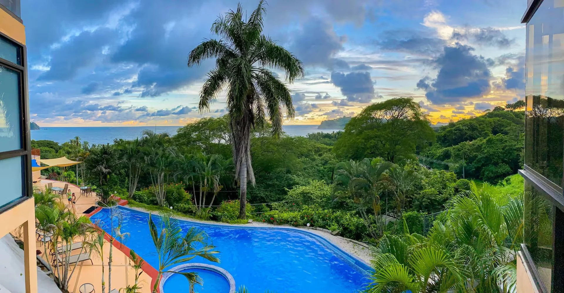 View from a balcony overlooking a lush tropical landscape with a large swimming pool, palm trees, and the ocean in the distance. The sky has clouds with the sun setting or rising.