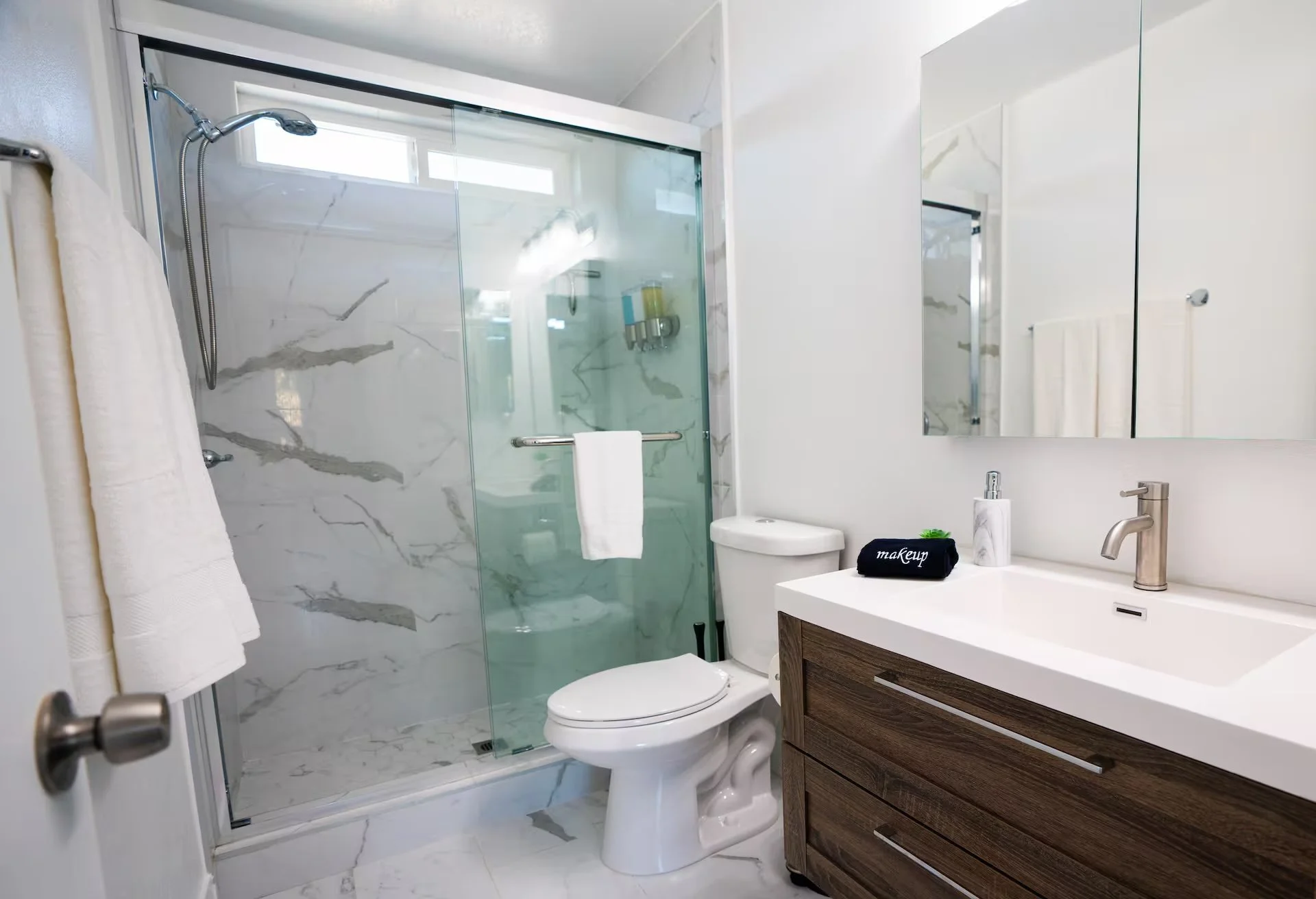 Modern bathroom with a glass-enclosed shower, white toilet, and a wooden vanity with a white sink, mirror, and soap dispenser.