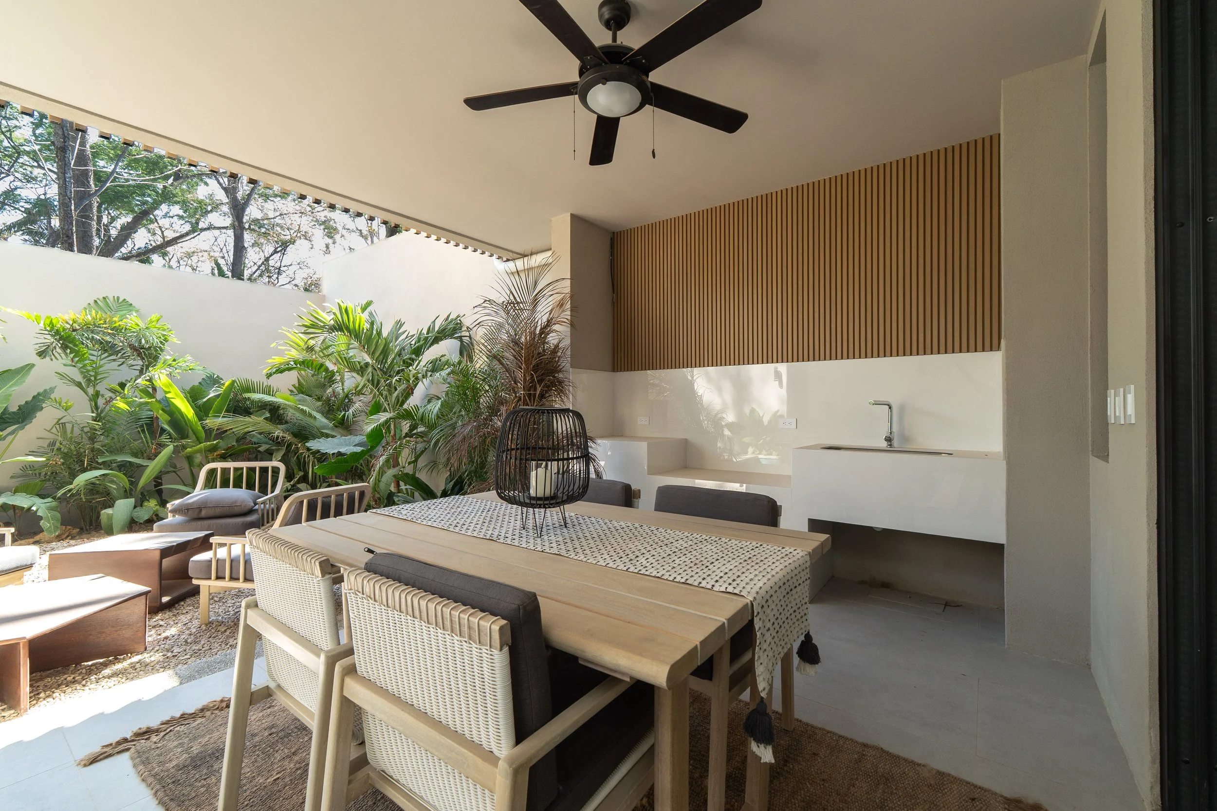 Outdoor patio area with a dining table, chairs, lush green plants, and a ceiling fan, partially covered by a roof.