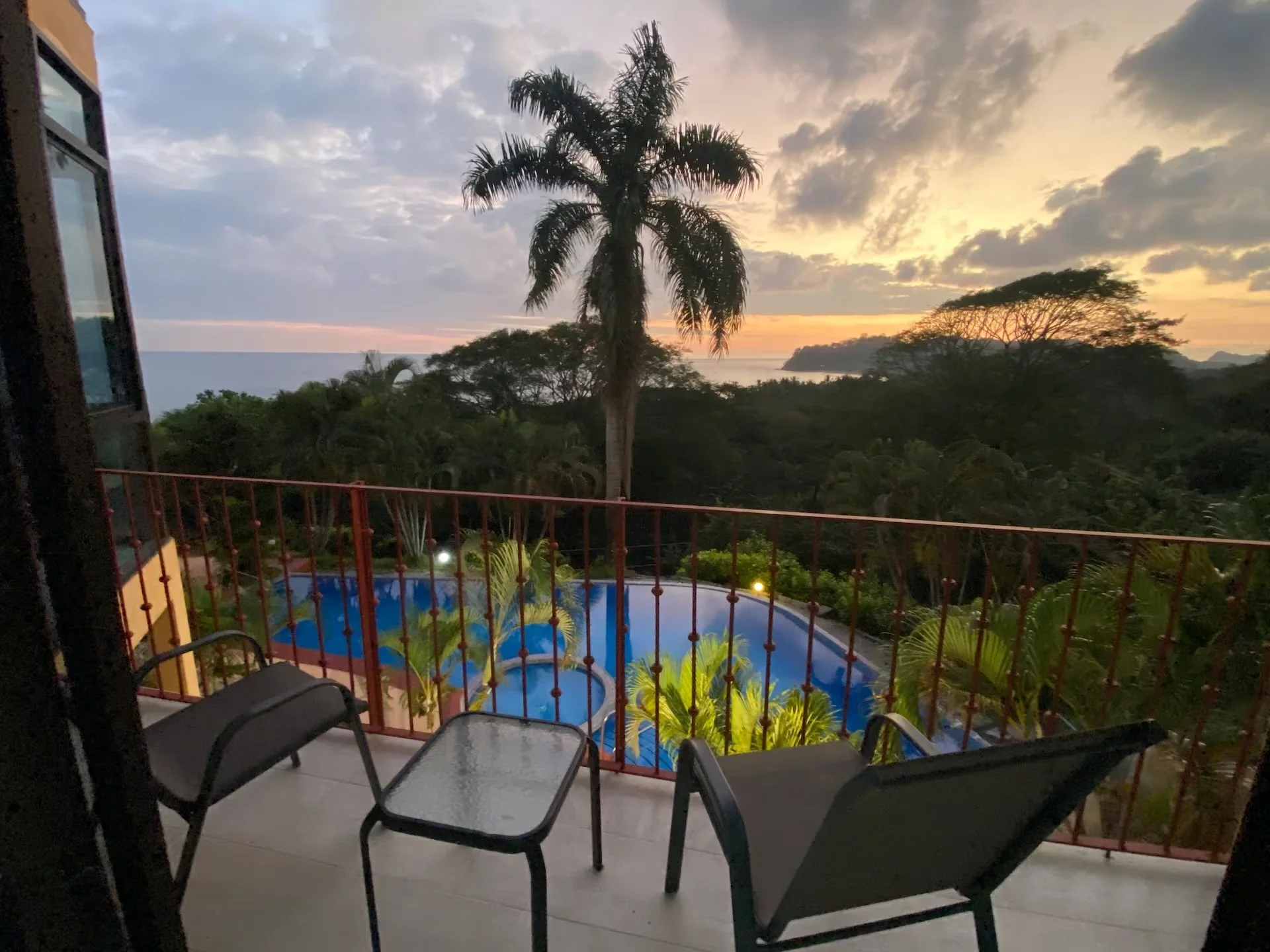 A balcony with two black chairs and a small glass table overlooking a lush tropical landscape with palm trees, a swimming pool, and a distant shoreline at sunset.