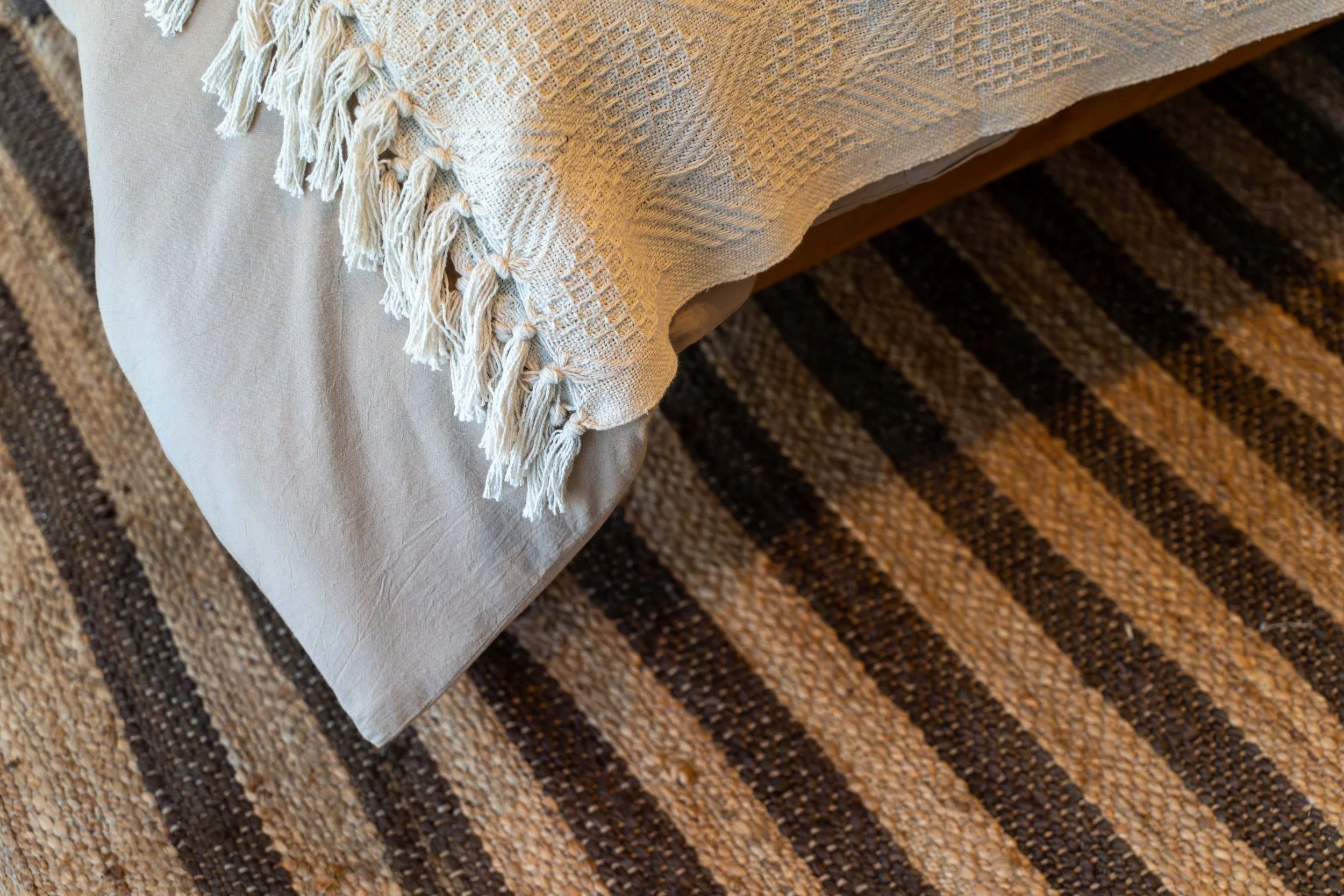 Close-up of a beige tablecloth with tassels on a wooden table, with a multicolored woven rug underneath.