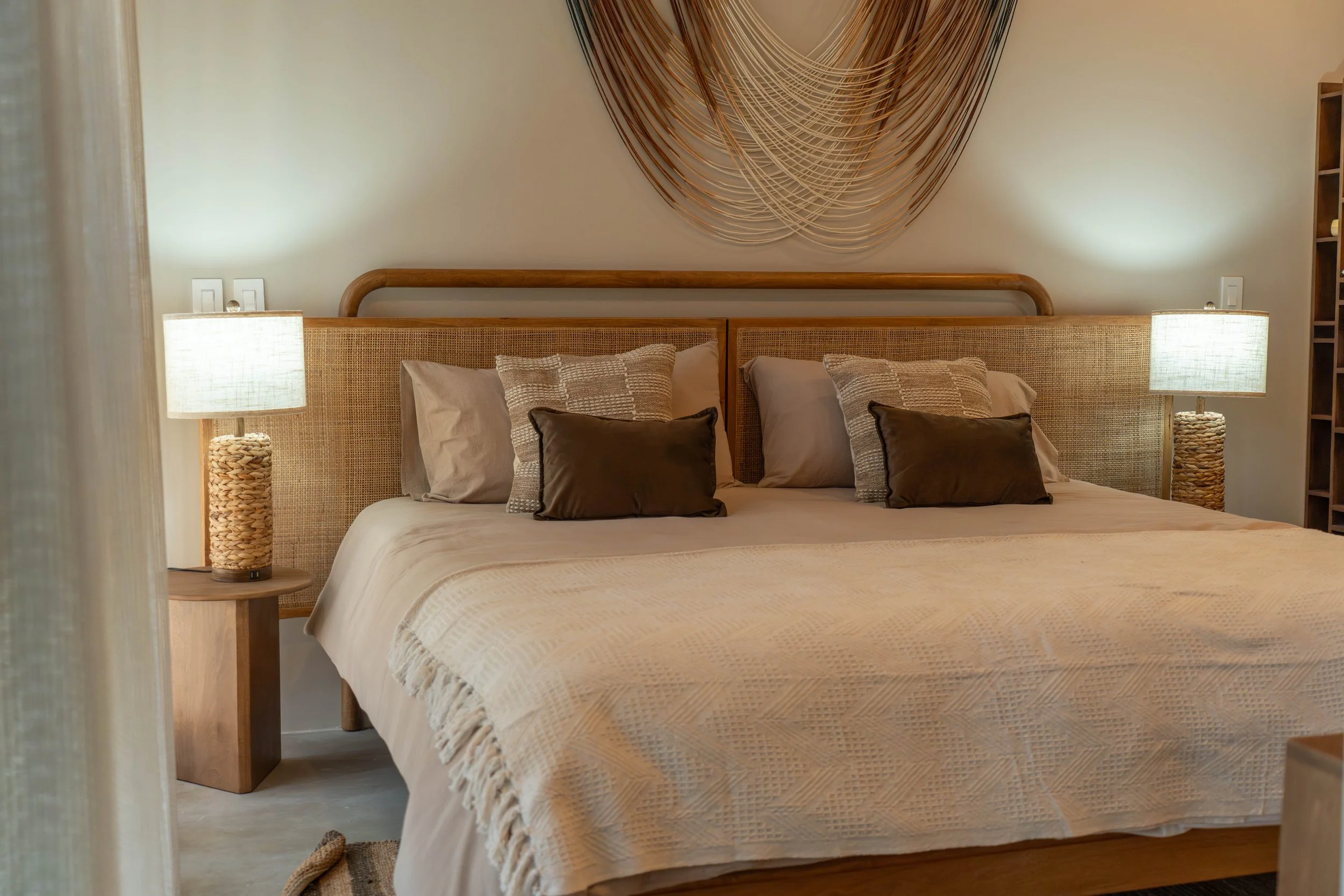 A neatly made bed with beige bedding and multiple brown, beige, and patterned pillows in a cozy bedroom. Two matching table lamps with woven bases sit on bedside tables on either side of the bed. A modern wall art piece made of curved wood strips han