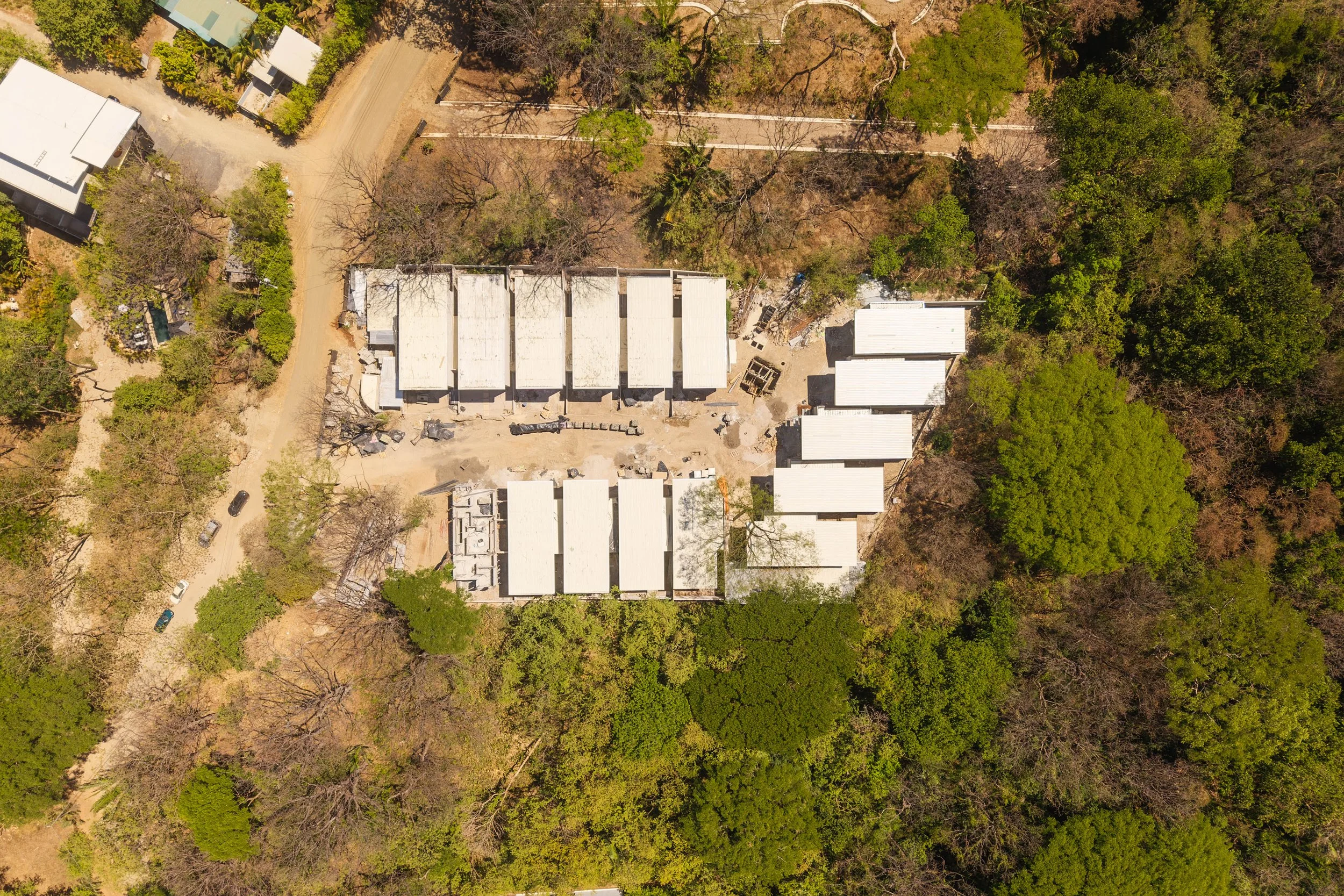 An aerial view of a residential area with multiple white-roofed buildings surrounded by trees, dirt roads, and green foliage.