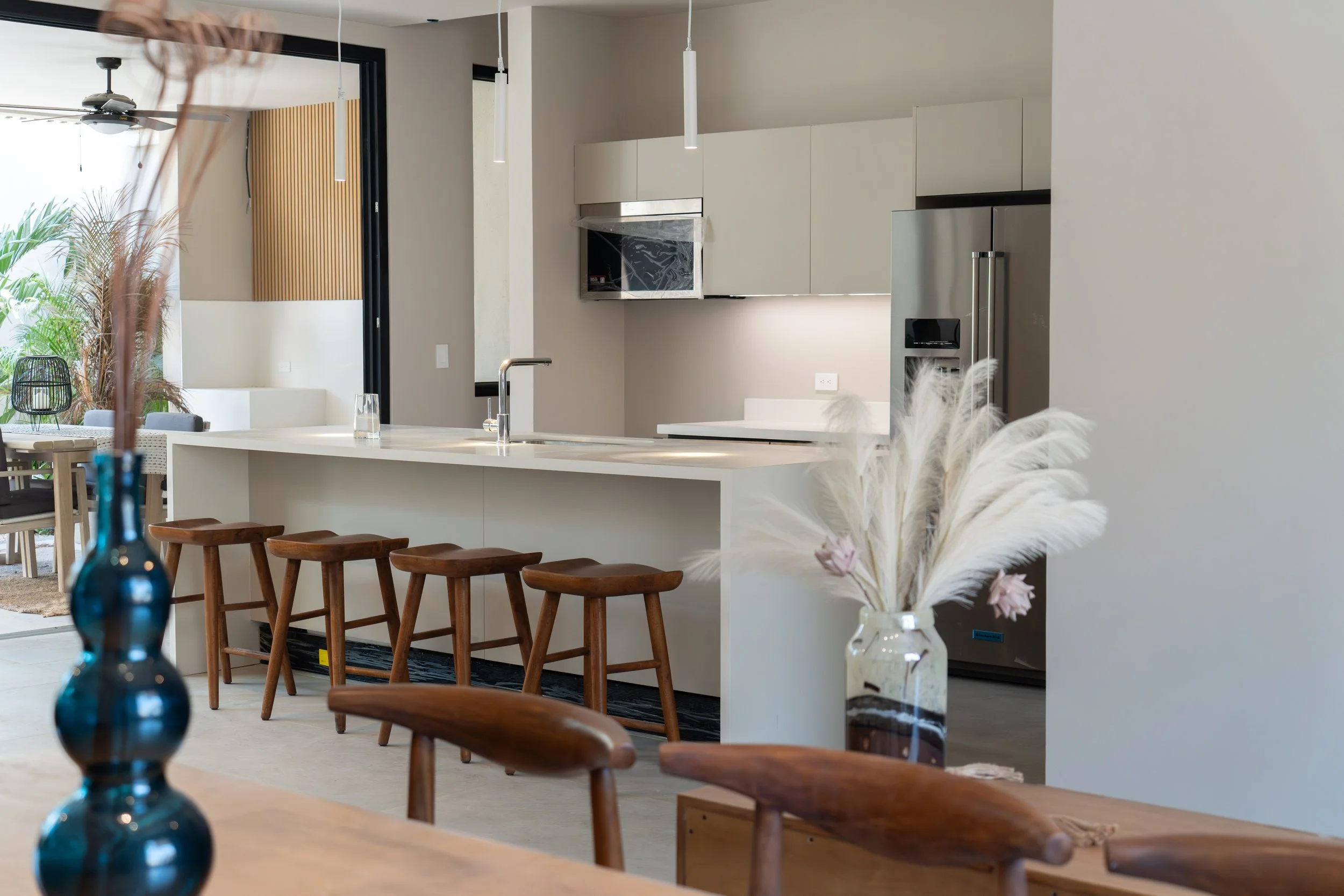 Modern kitchen with white cabinetry, stainless steel appliances, and a large island with a faucet. There are wooden barstools at the island, pendant lights hanging from the ceiling, and a view into a dining area and living room with a ceiling fan and