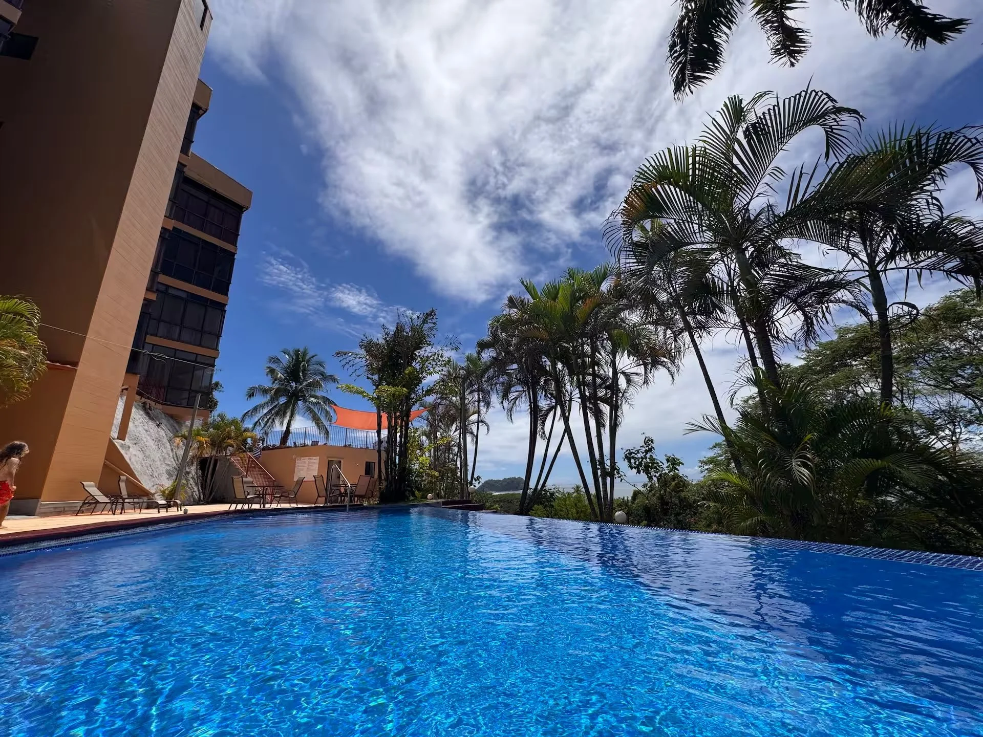 A swimming pool surrounded by palm trees and a clear blue sky with some scattered clouds, adjacent to a multi-story building with balconies and lounge chairs.
