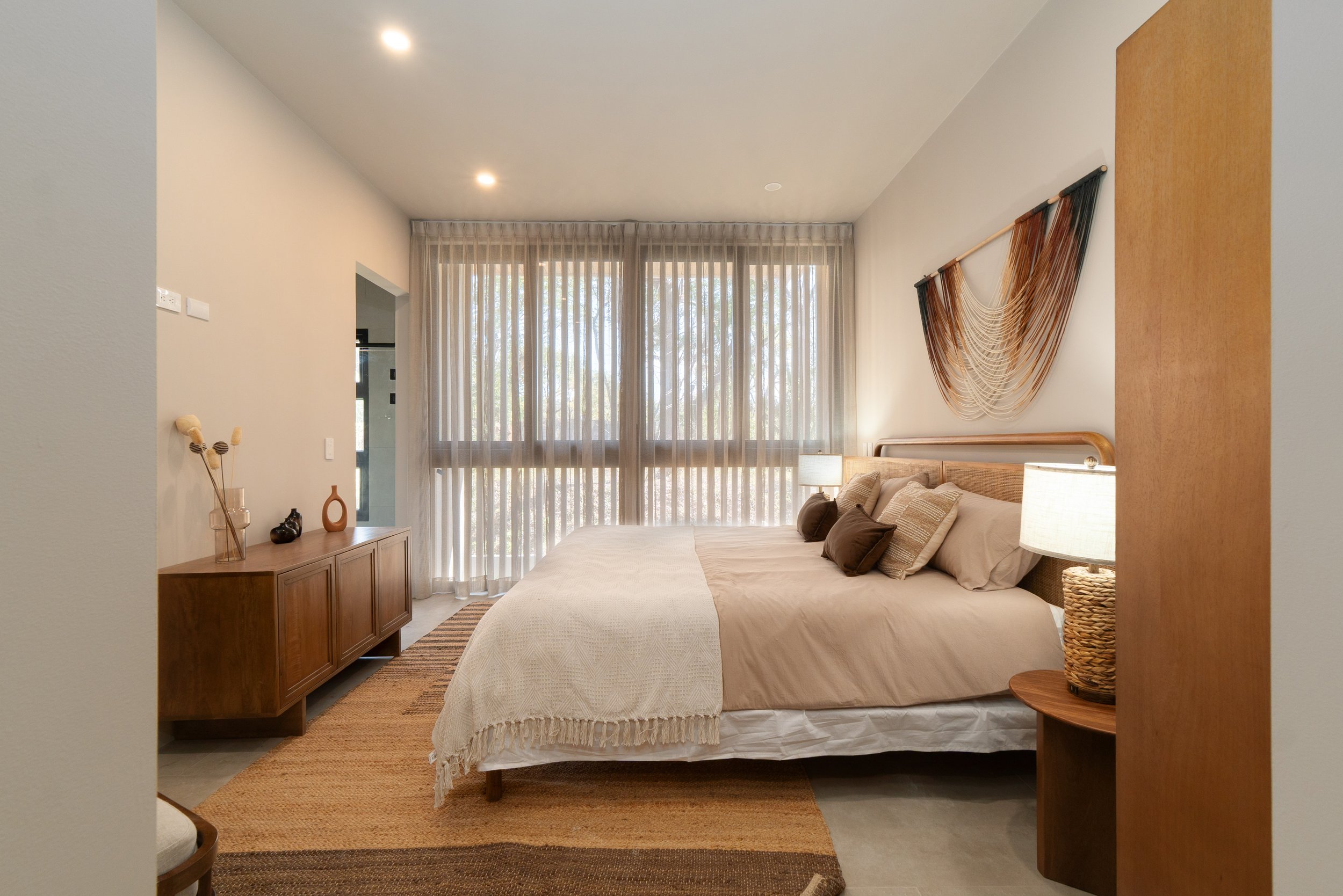 A bedroom with a large bed, nightstands, and decorative wall hanging, with sheer curtains and large windows letting in natural light.