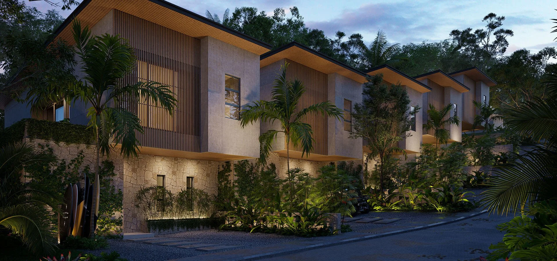Modern multi-story residential building with large windows, surrounded by lush tropical plants and trees, illuminated at dusk.