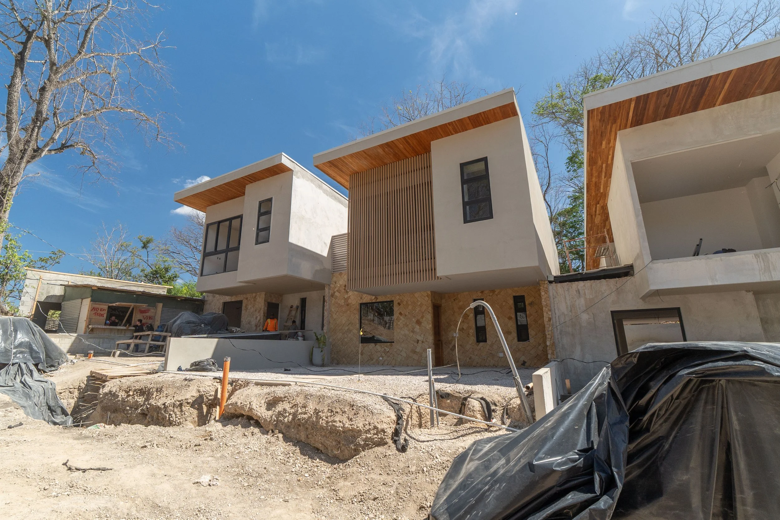 Modern multi-story residential building under construction with white walls, wooden accents, and large windows, set against a clear blue sky, with construction materials and debris in the foreground.