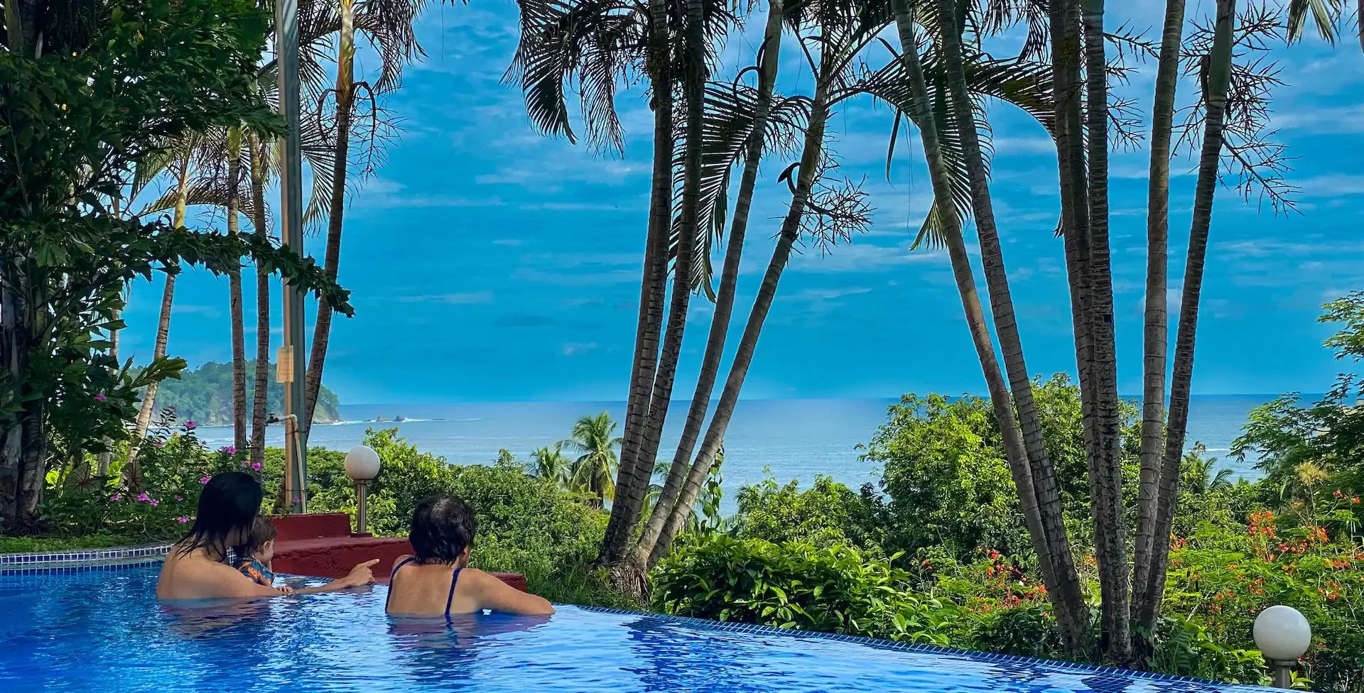 Three people in a pool overlooking a tropical landscape with lush trees, palm trees, and the ocean in the background under a partly cloudy sky.