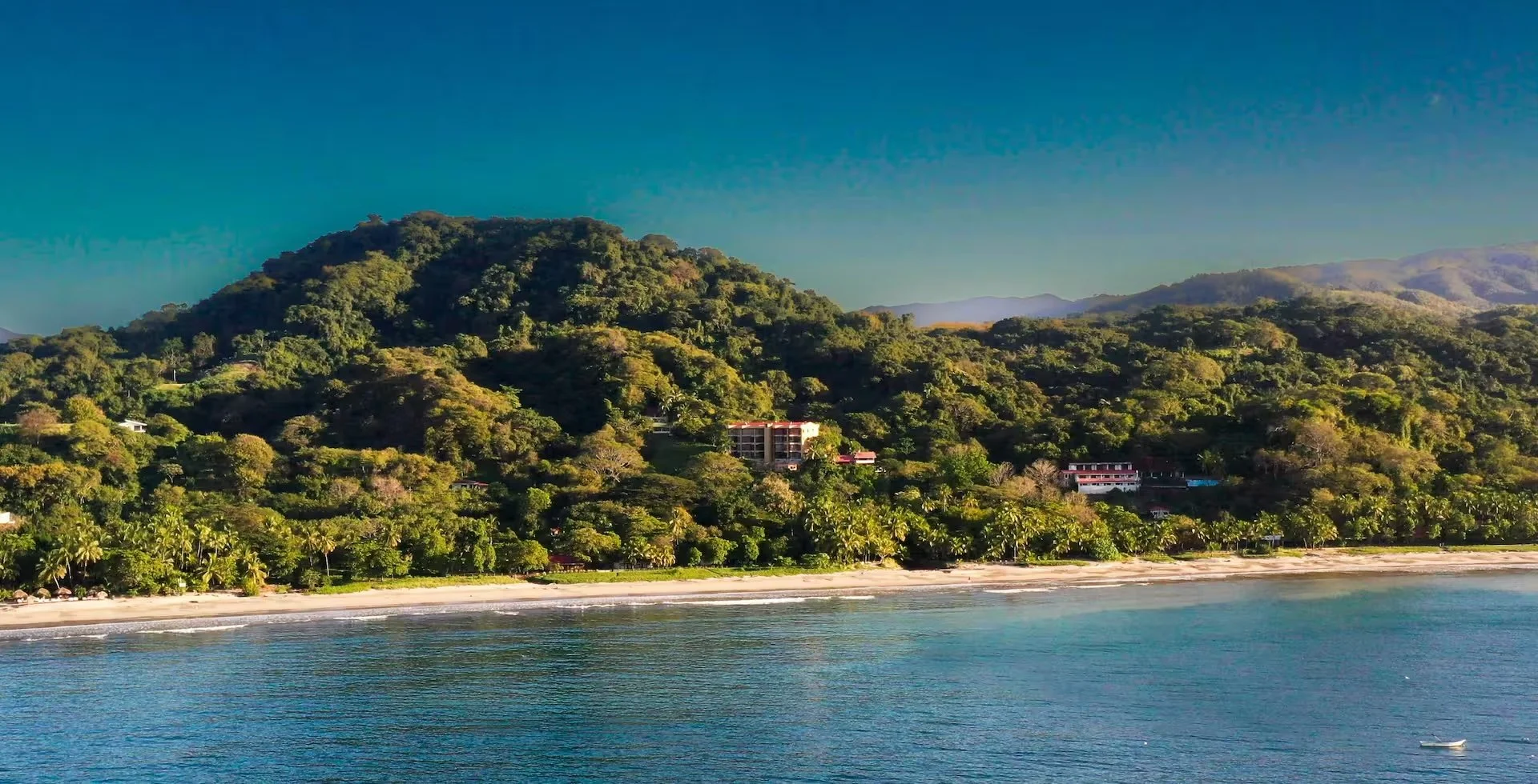 Tropical beach with calm blue water, sandy shoreline, lush green trees and hills in the background under a clear blue sky.