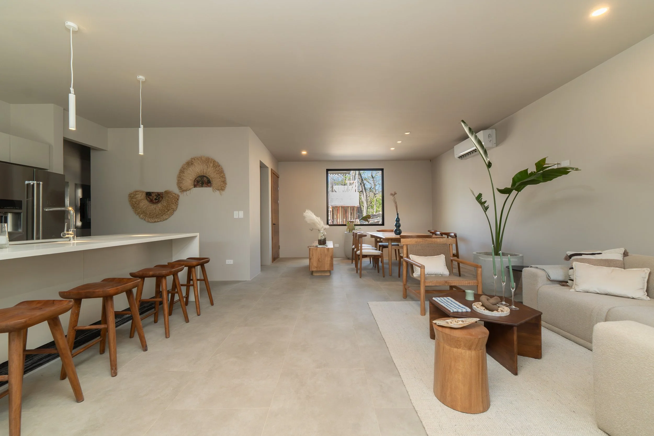 Open-concept living and dining area with modern furniture, a large green plant, wall decorations, and natural light from window, with neutral color palette.