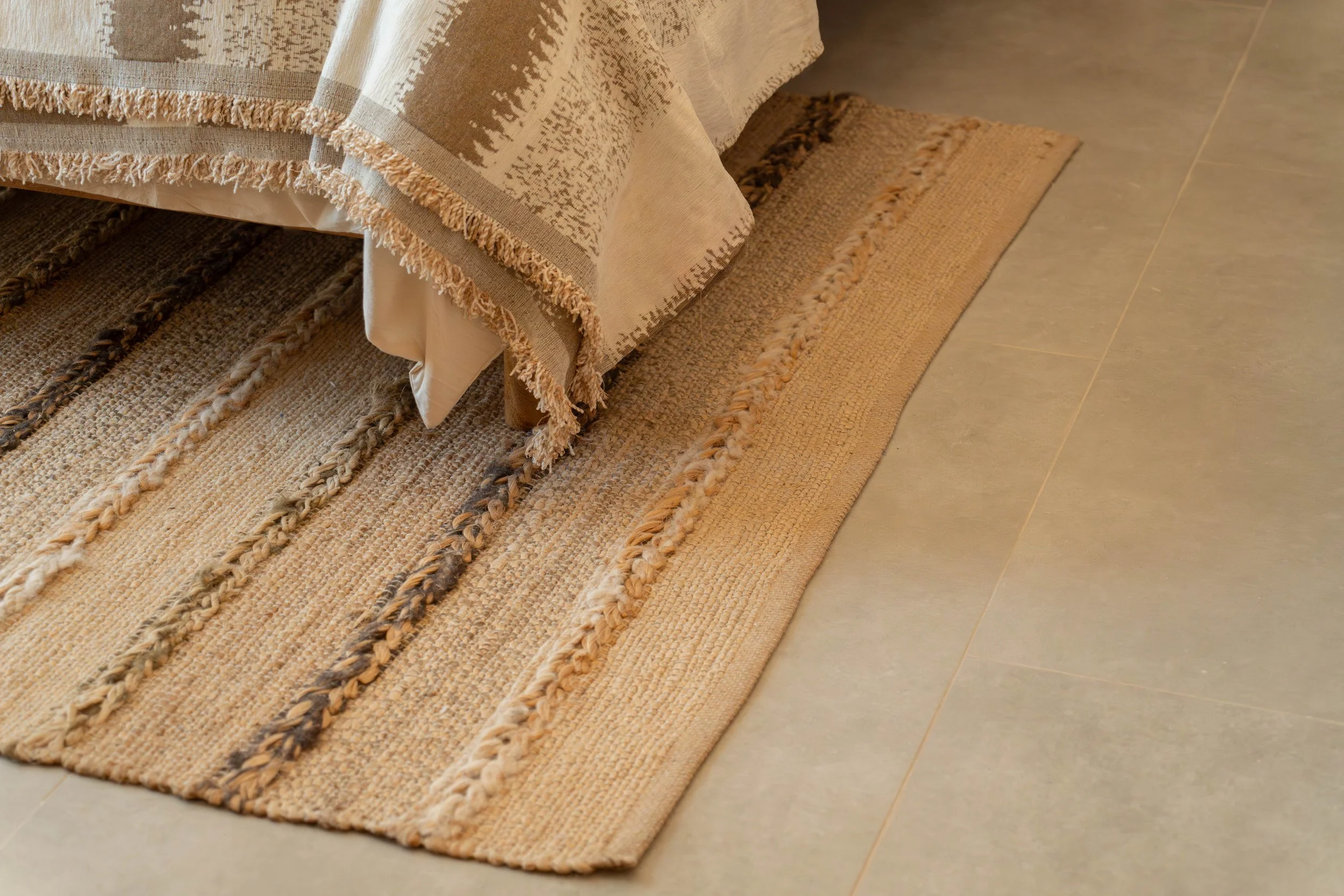 Close-up of a woven beige and brown area rug layered on top of a larger rug, next to a tiled floor. The layered rugs feature textured, braided designs and fringed edges, with part of a fabric-covered piece of furniture visible.