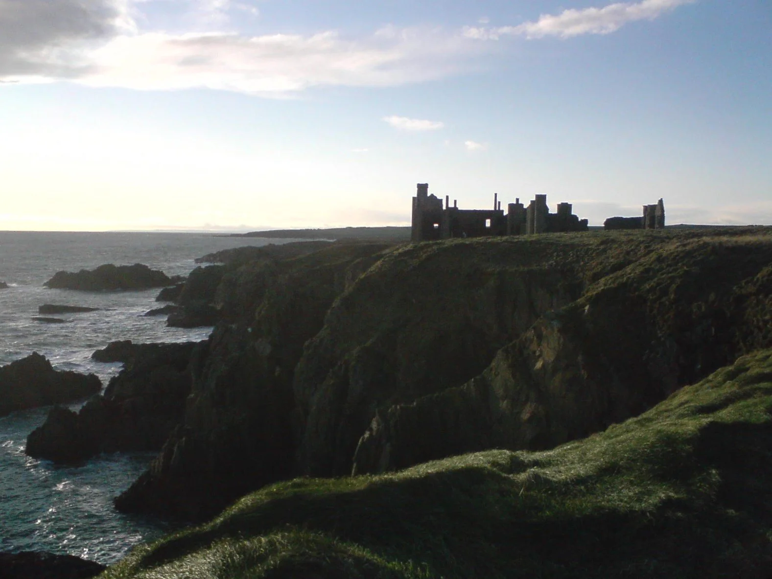 Ruins of a castle on a grassy cliff overlooking the ocean at sunset or sunrise.