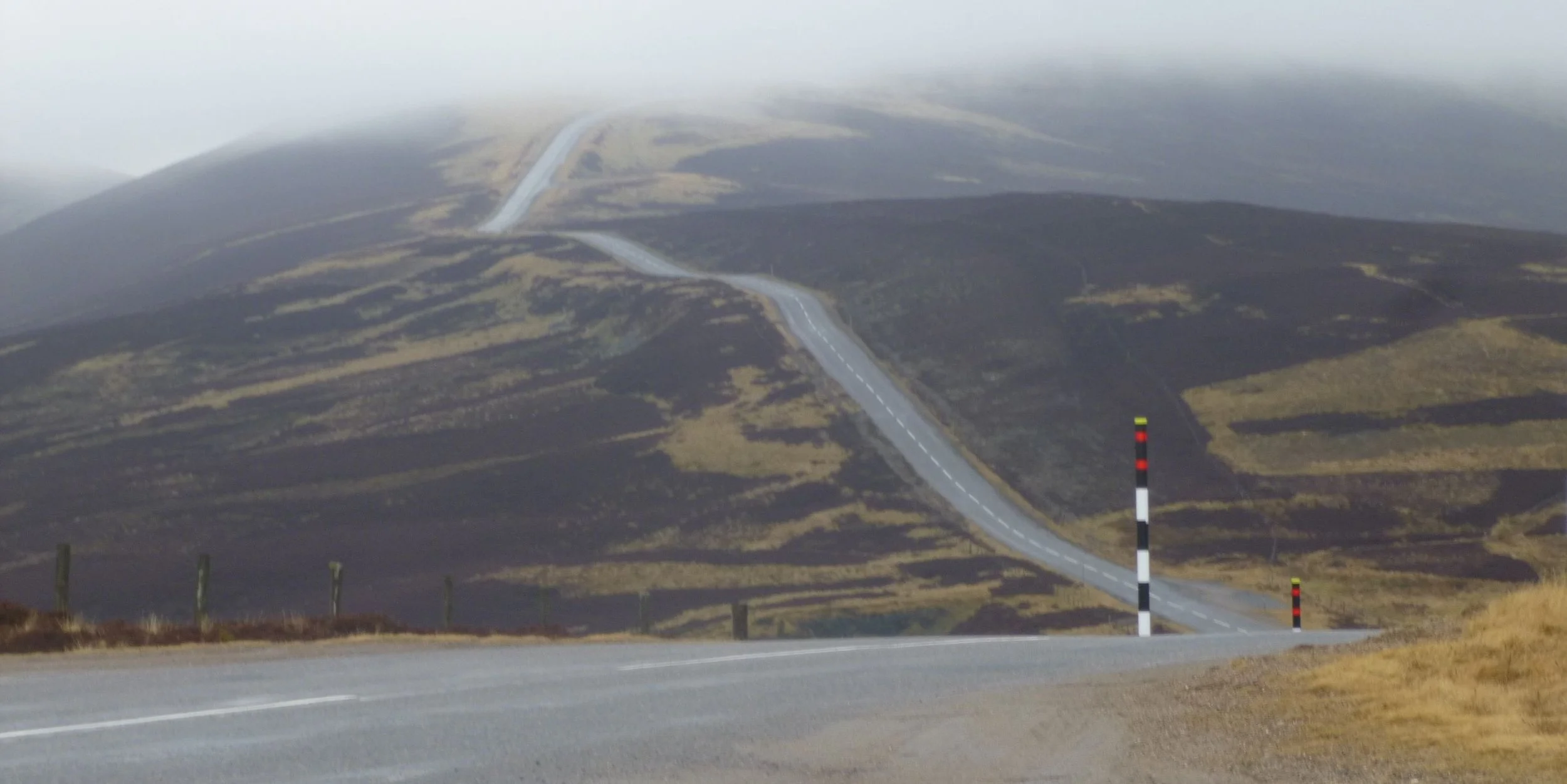 A winding mountain road with black and white striped posts on the side, going uphill through a mountainous landscape with grassy patches and a misty, foggy sky.