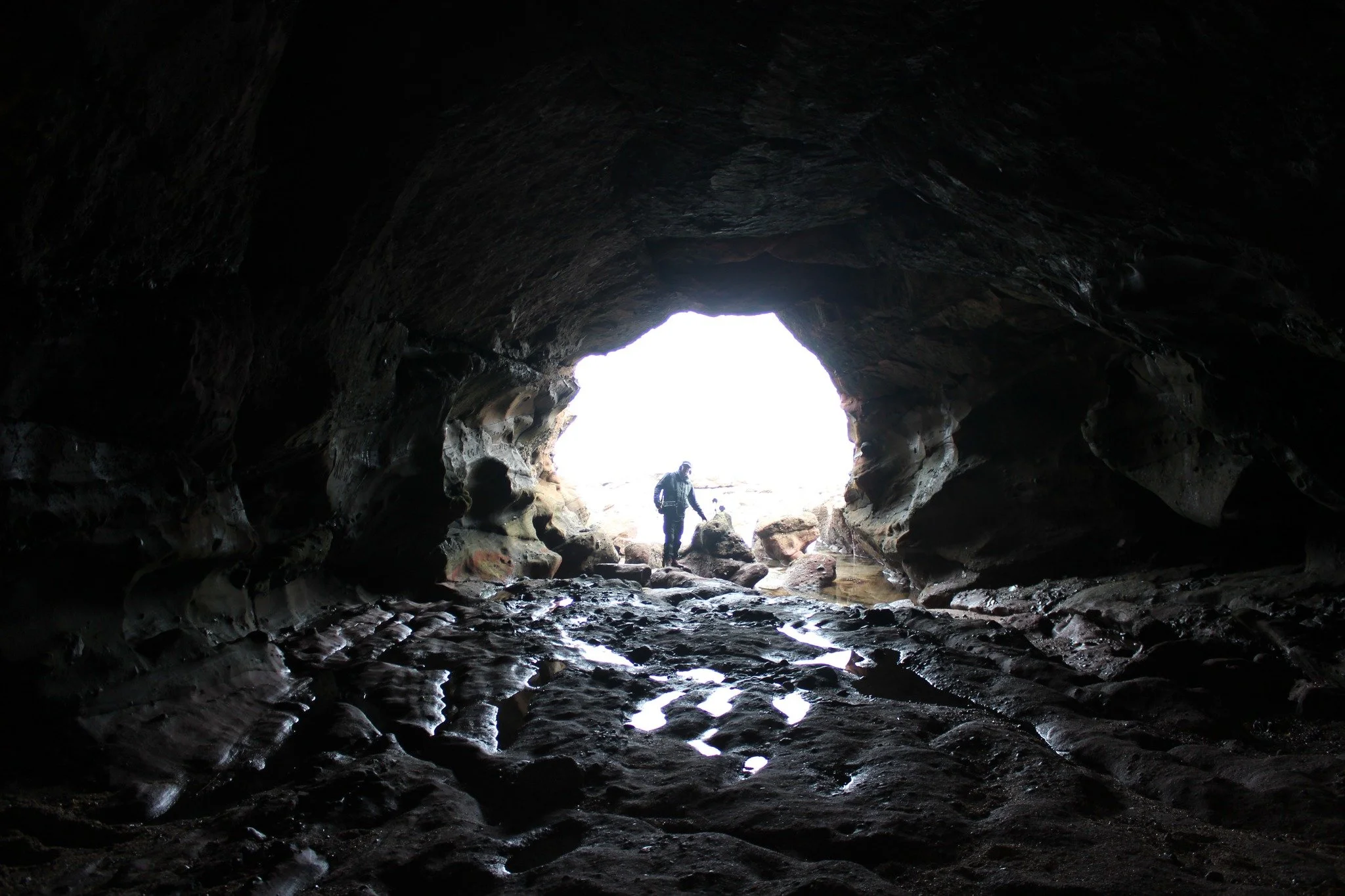 A person exploring a dark cave with an opening showing bright outdoor light and rocks around