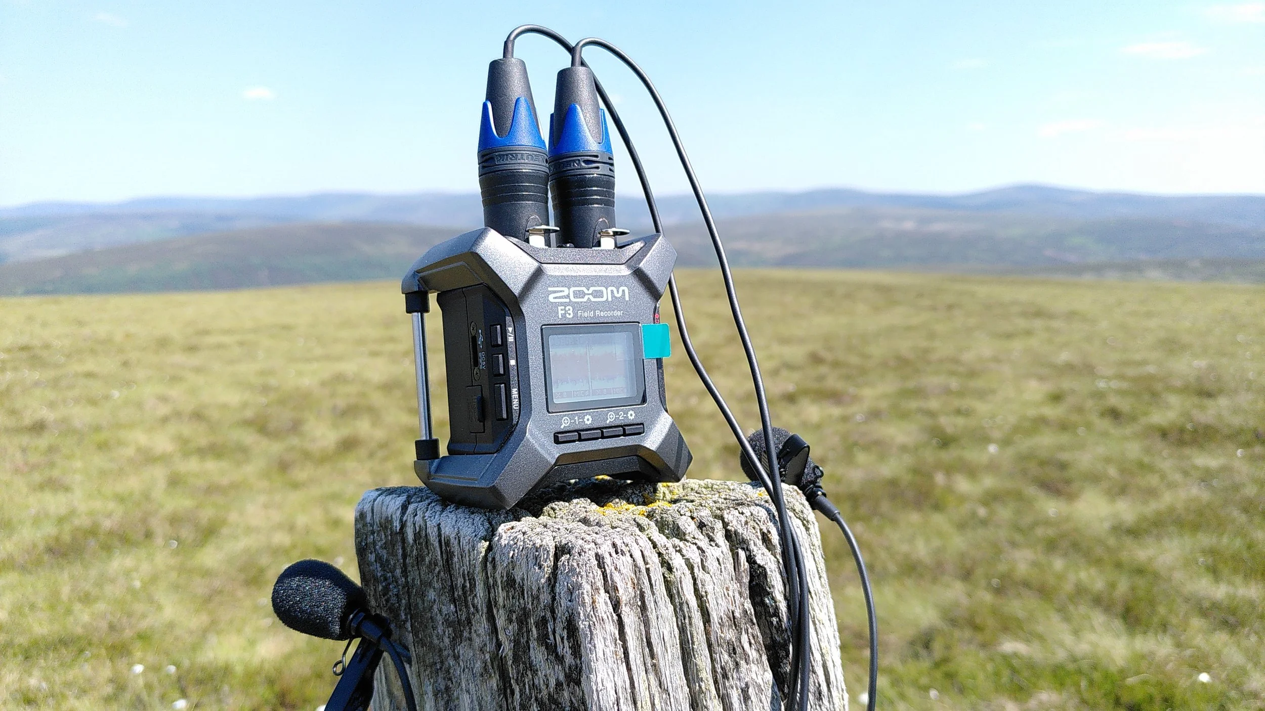 A portable audio recorder with microphones set up outdoors on a weathered wooden post in a grassy field, with mountains and a blue sky in the distance.