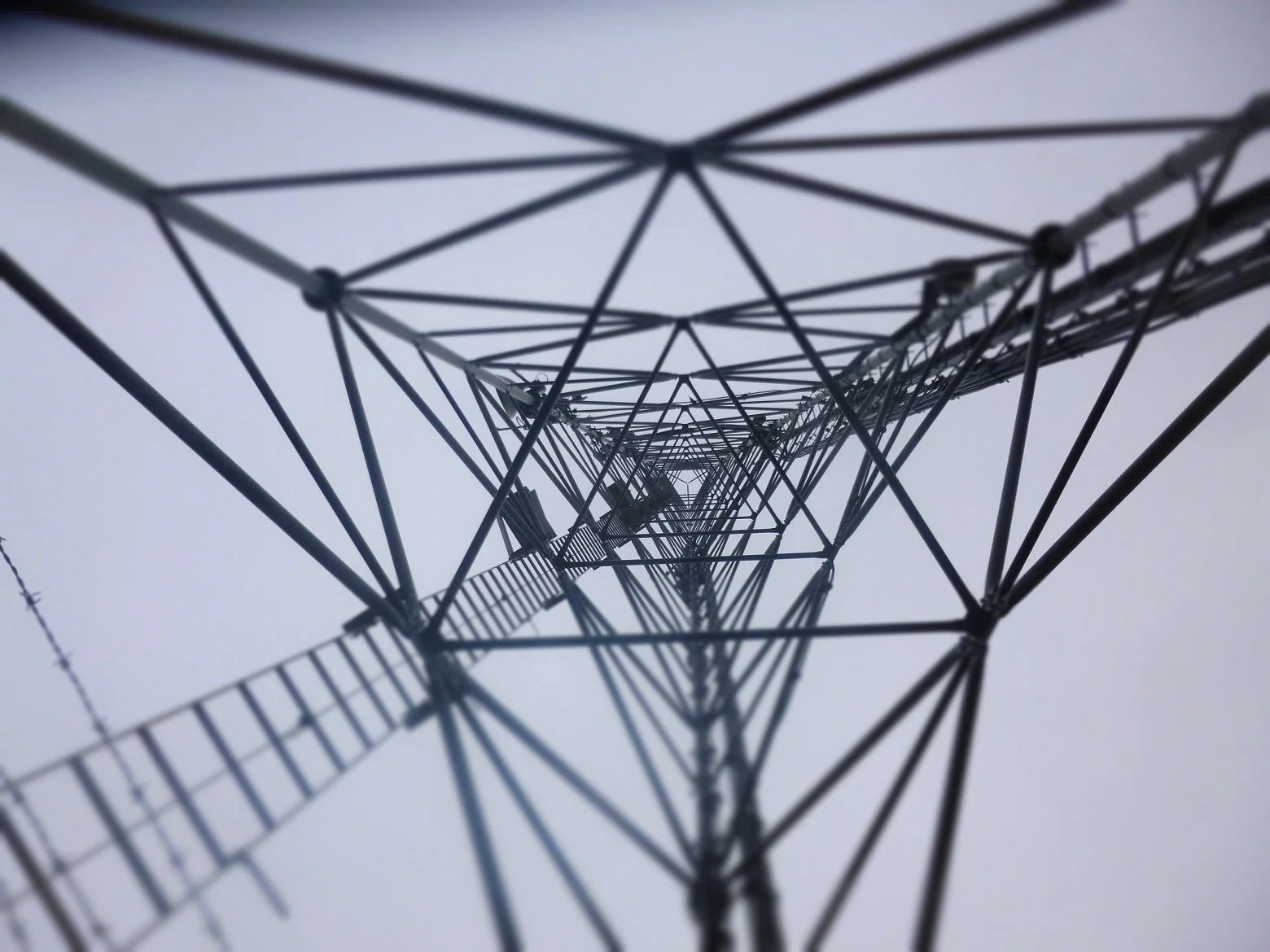 Looking up at a tall radio tower with a ladder running along its side, against a gray sky.