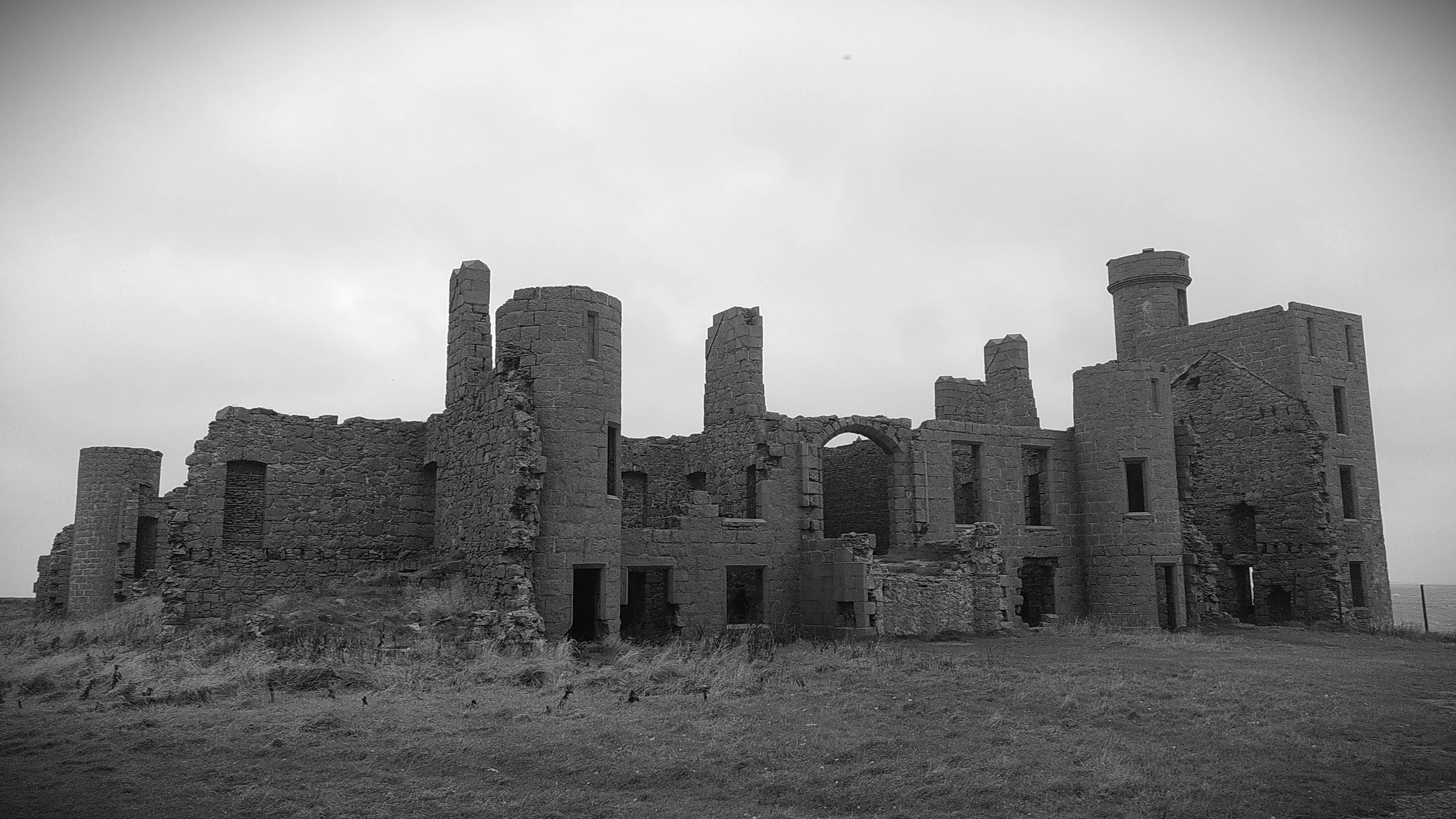 Black-and-white photo of the ruins of a stone castle with multiple towers and broken walls, situated on grassy terrain under cloudy sky.