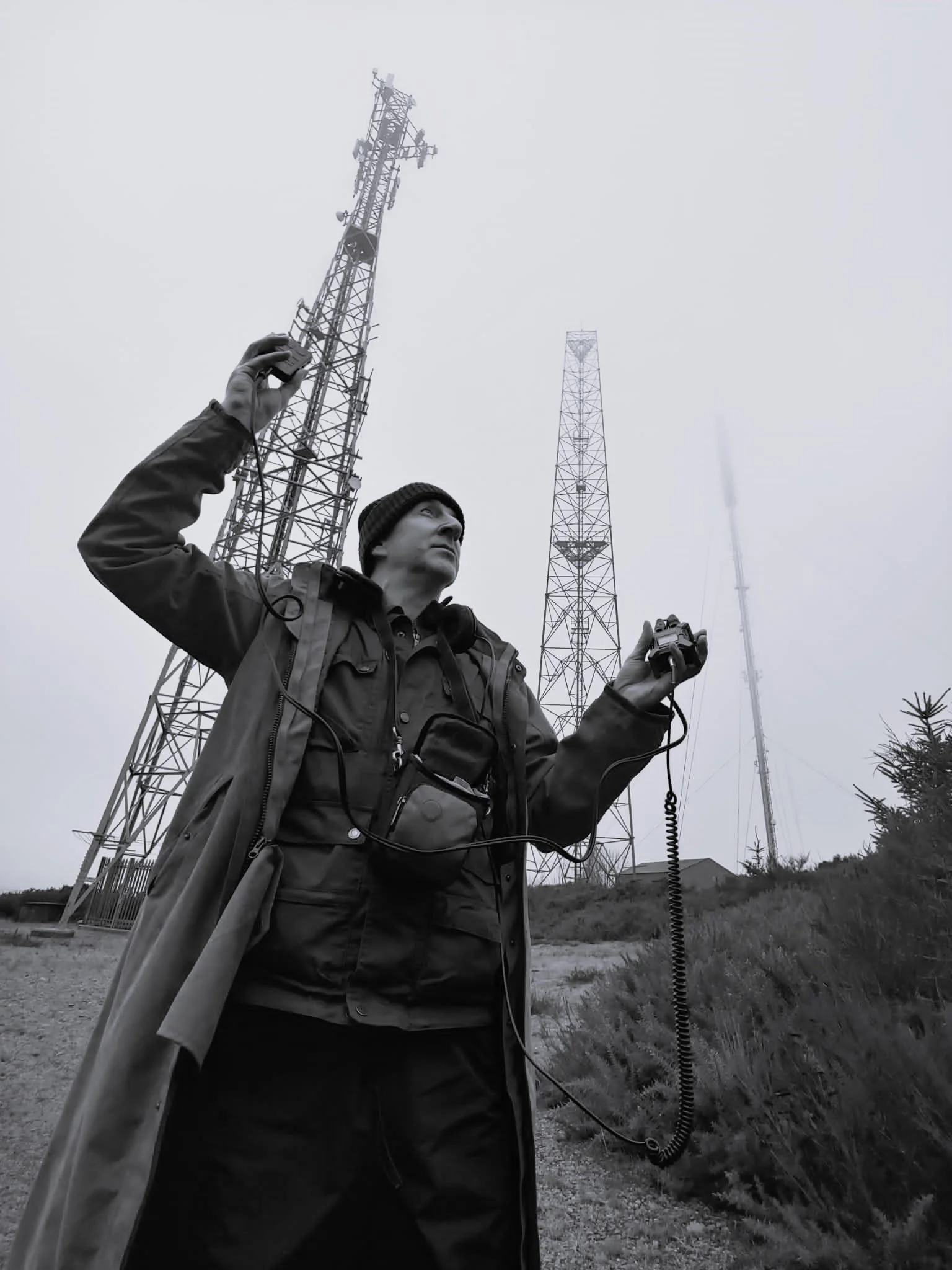 A man outdoors in front of communication towers, holding a radio device and a microphone.