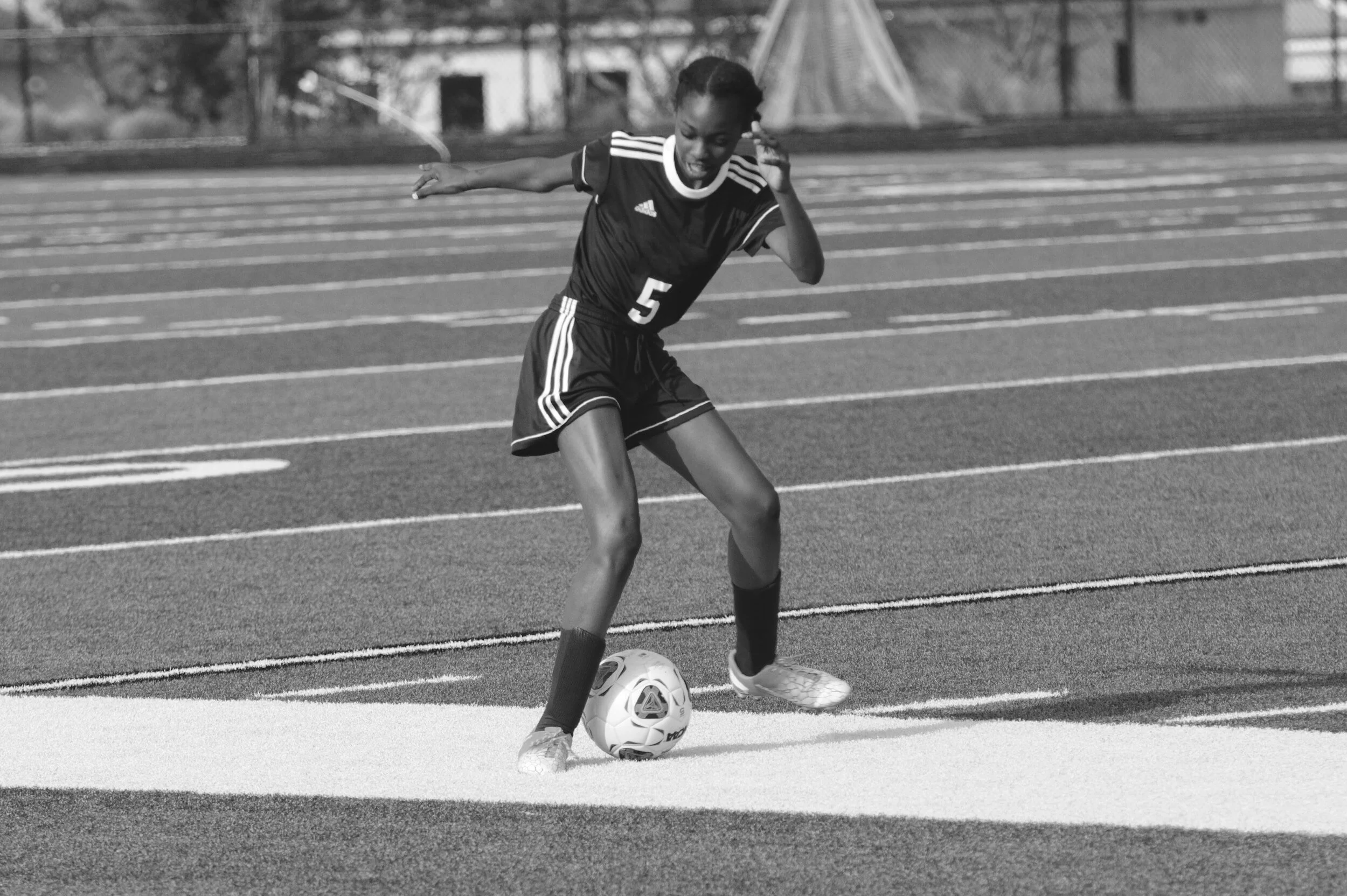A female soccer player in a black uniform with the number 5 on her jersey, standing on a football field with lines, preparing to kick a soccer ball.
