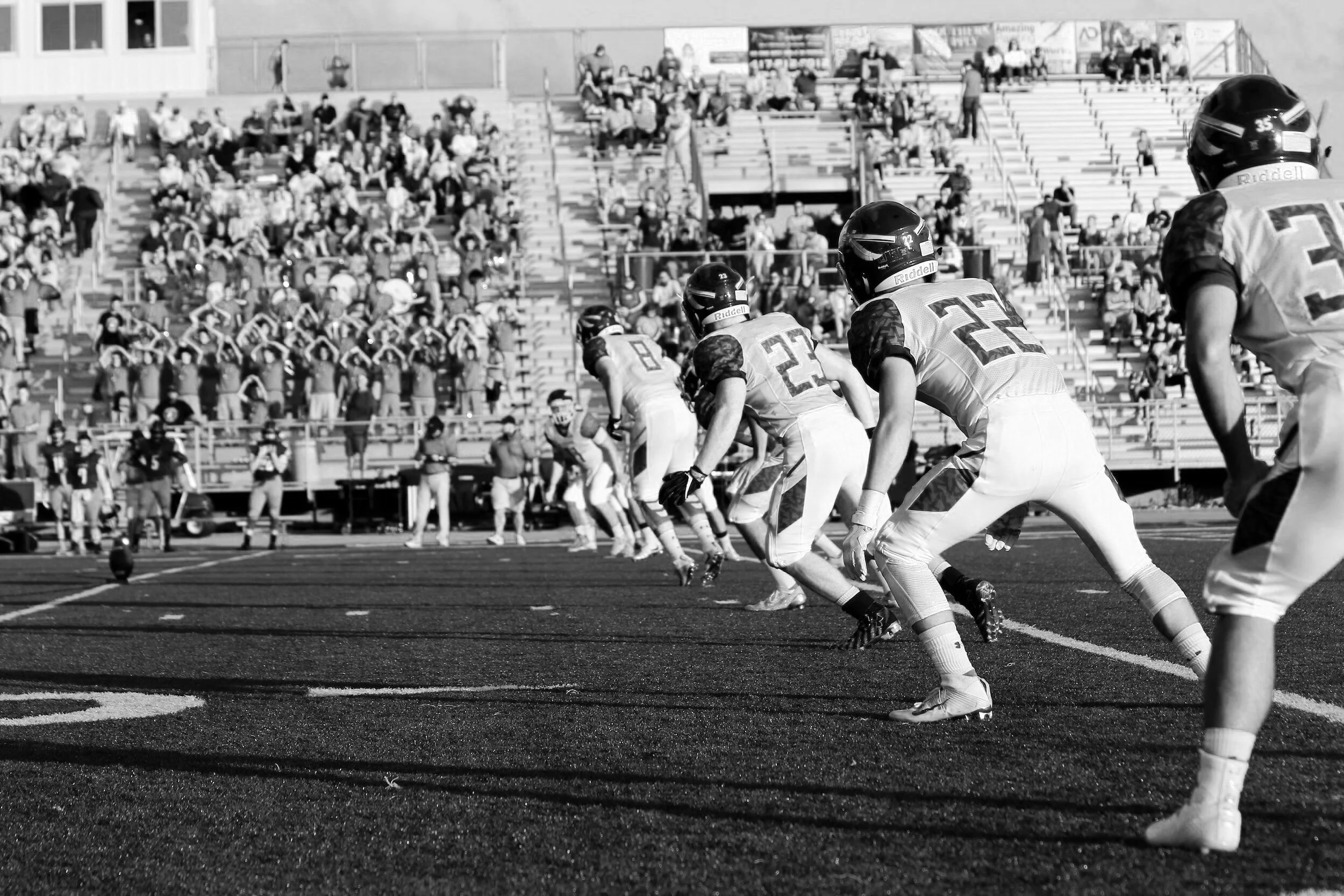 American football players lined up at the start of a game on a football field, with a crowd of spectators in the stands behind them.