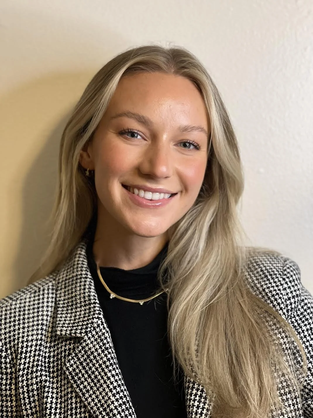 A young woman with long blonde hair smiling, wearing a black top and patterned blazer, against a plain light-colored wall.