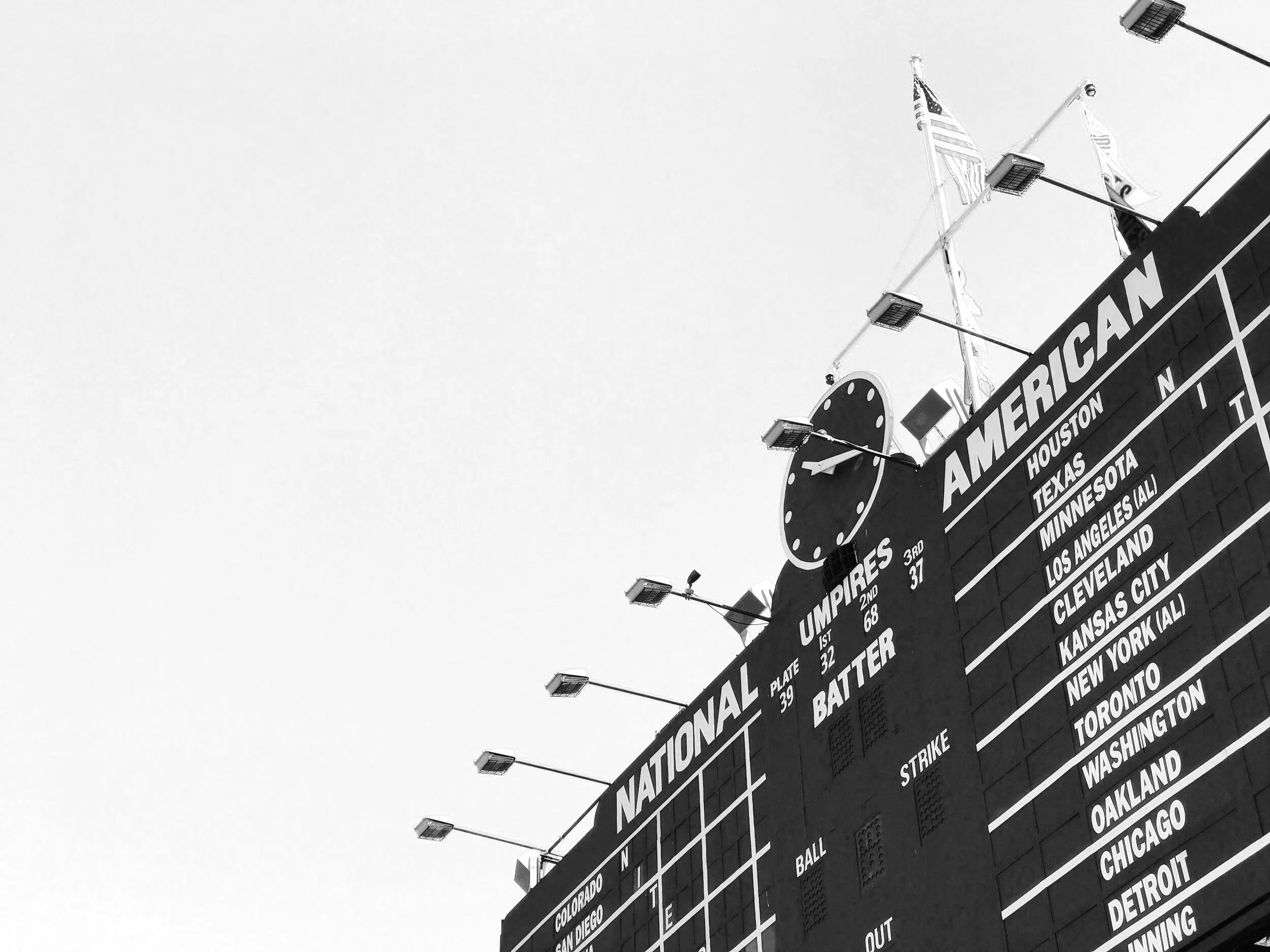 Part of a baseball scoreboard at a stadium, showing team names and rankings, with flags and lights above.