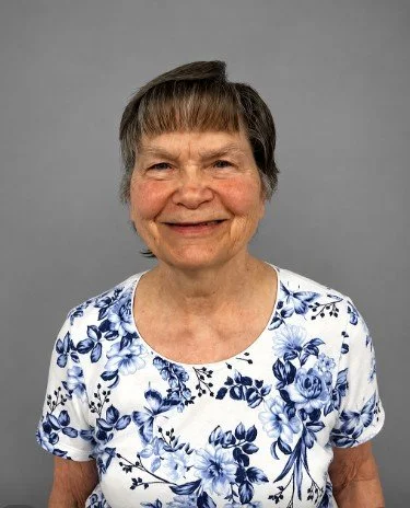 An elderly woman with short brown hair and a smile, wearing a white shirt with blue floral pattern, standing against a plain white wall.
