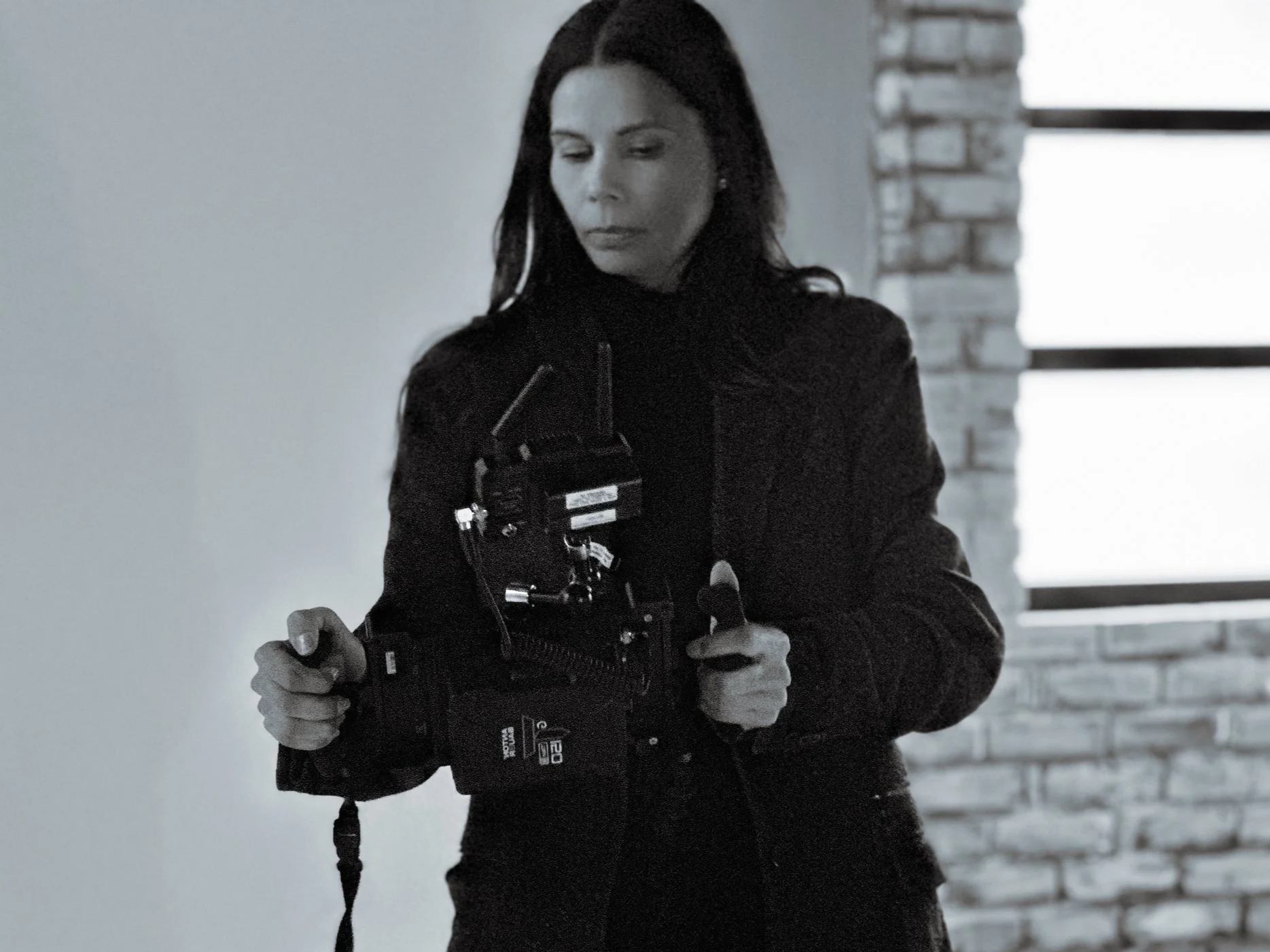 Regina Dezbah Simons holds a monitor on set of her film, standing beside a brick wall and a large window in studio.