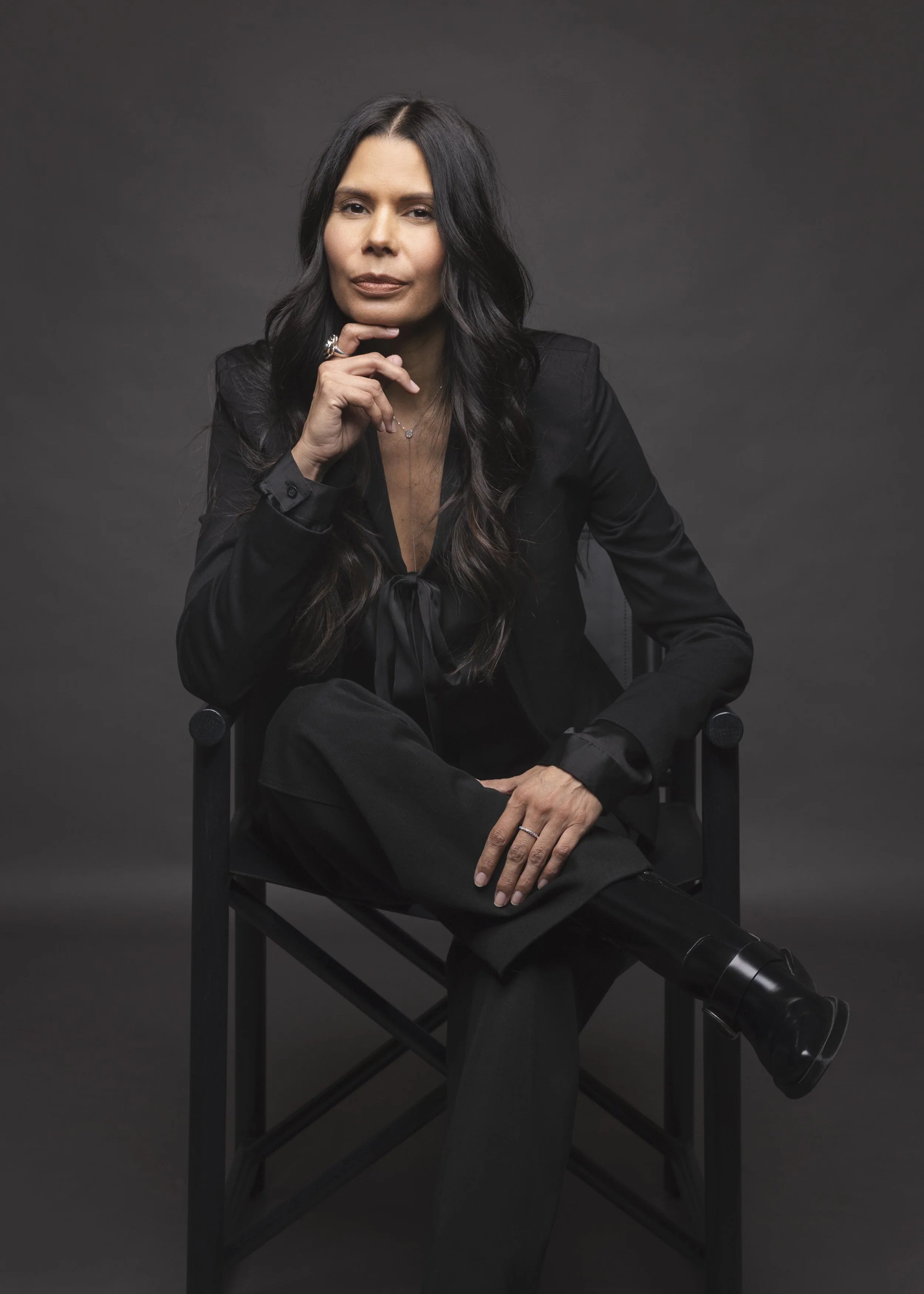 Regina Simons sitting on a director's chair, dressed in a black suit with long dark hair, posing in front of a dark background.