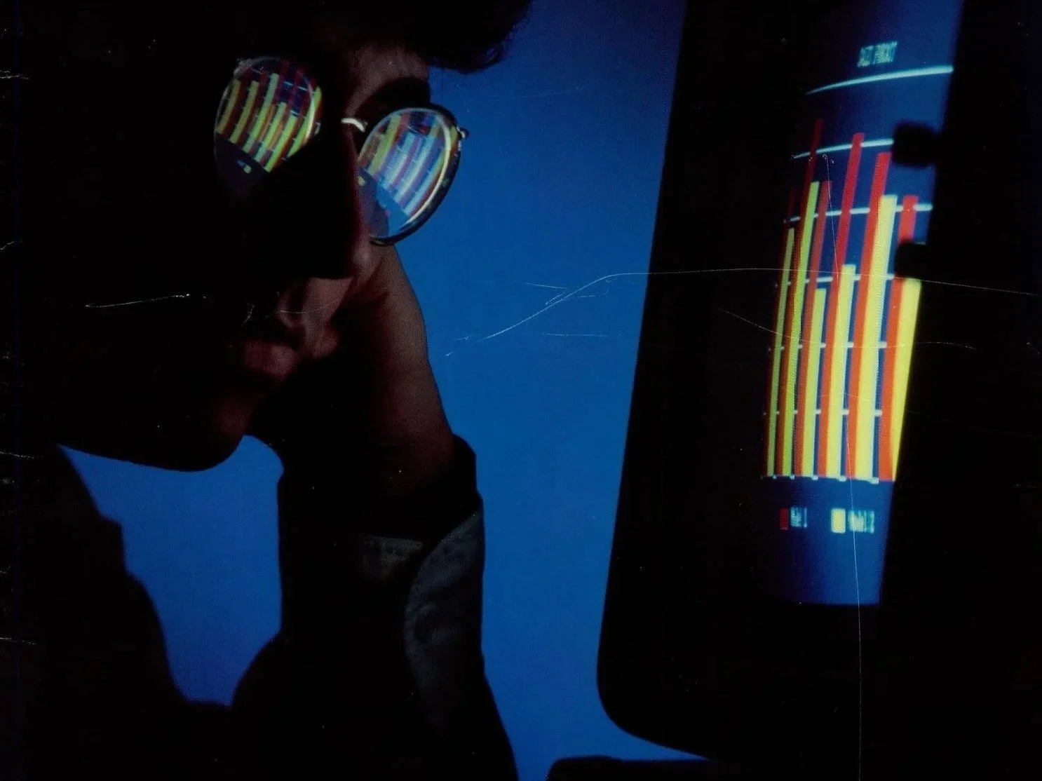 A person in shadow rests their chin on one hand and studies a red and yellow bar chart on a computer screen, the chart reflected in their round glasses against a deep blue background.