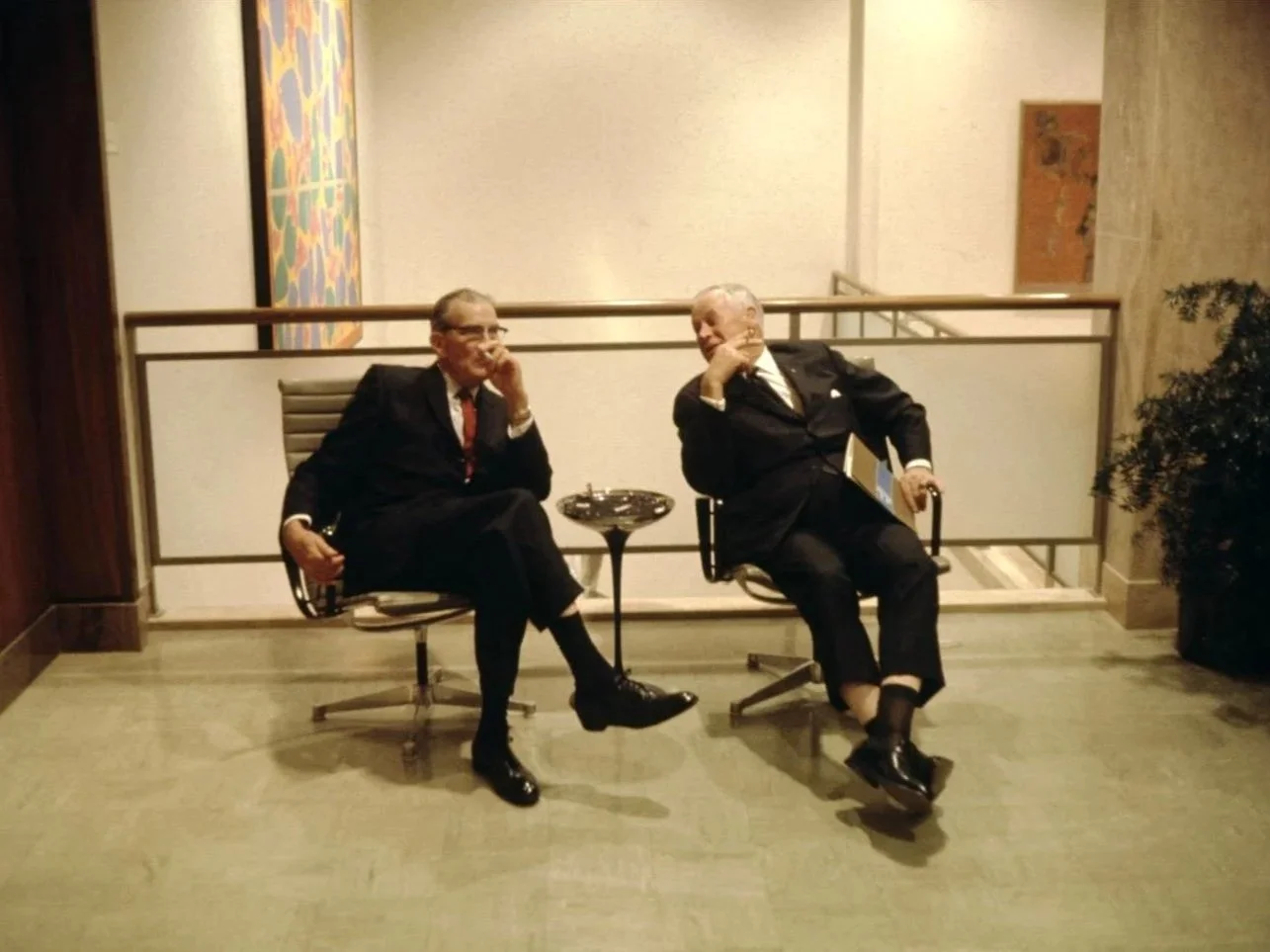 Two men in dark suits seated in Eames-style chairs in a modernist lobby, conversing, with an abstract painting visible on the wall behind them.