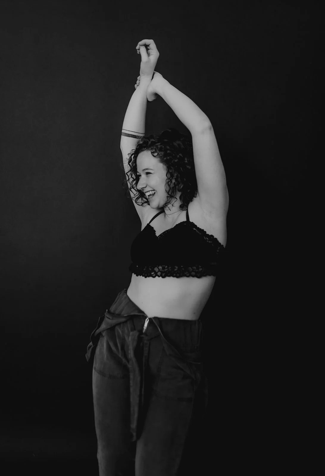 Photo of Savannah during a self love photo session. Arms up with her jump suit partially down in a bralette.