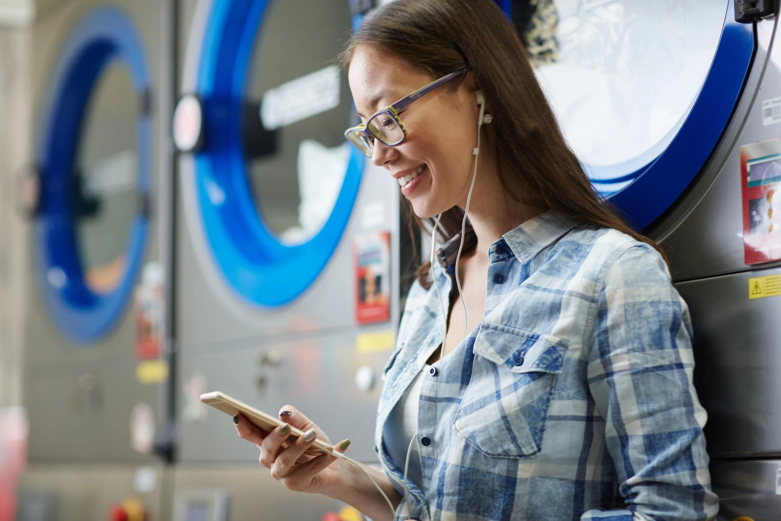 Asian woman smiling and looking down at her cellphone in front of laundromat dryers